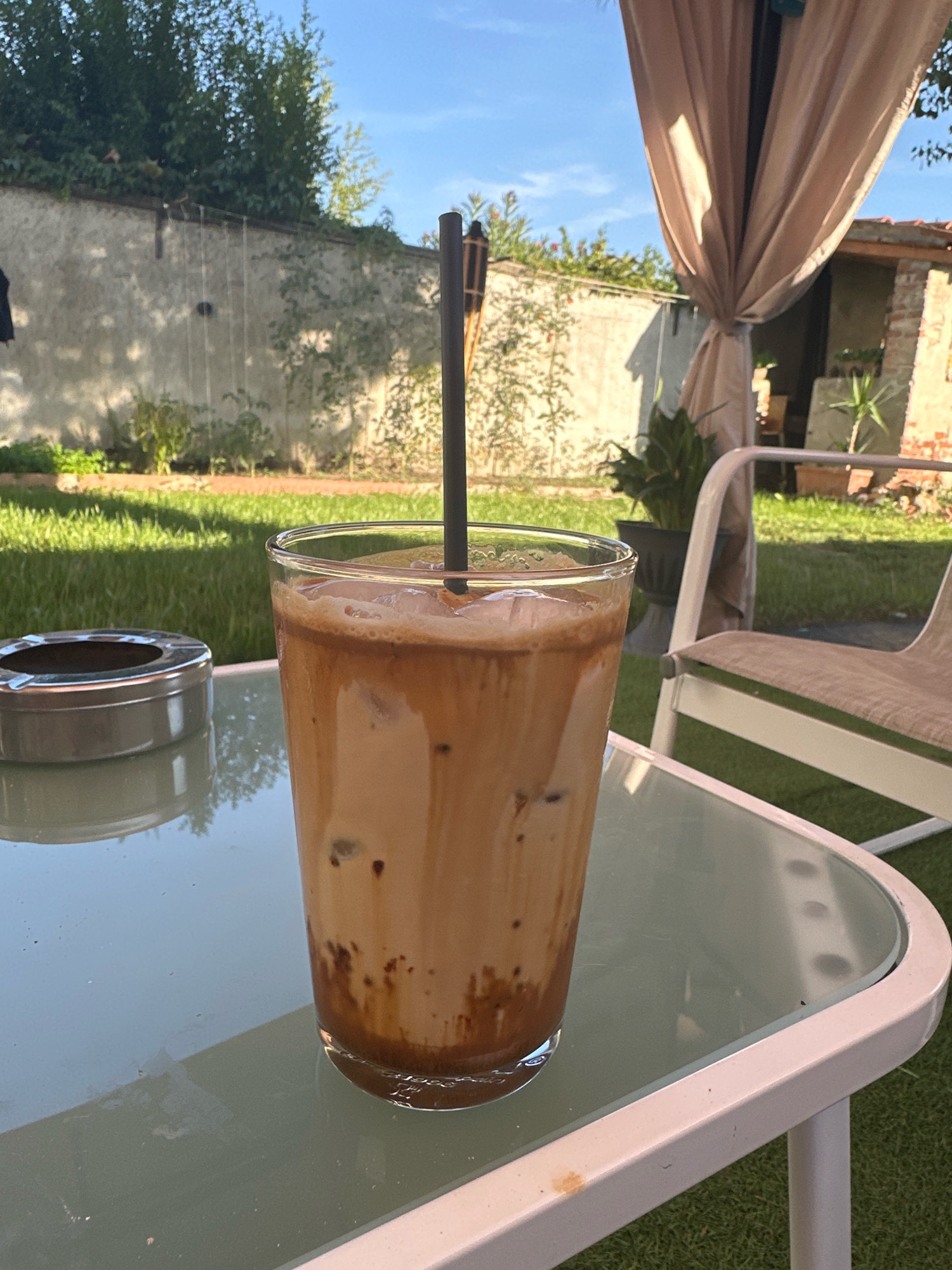 An iced coffee with a black straw sits on a glass-topped table in an outdoor setting. To the left of the coffee is a metal ashtray. In the background, a stone wall is partially covered with ivy and foliage, with a thatched-roof structure and a potted plant visible beyond. A beige awning hangs from the right side of the frame. The ground is covered in green grass. The scene appears to be in Villa Pasquali, Italy.
