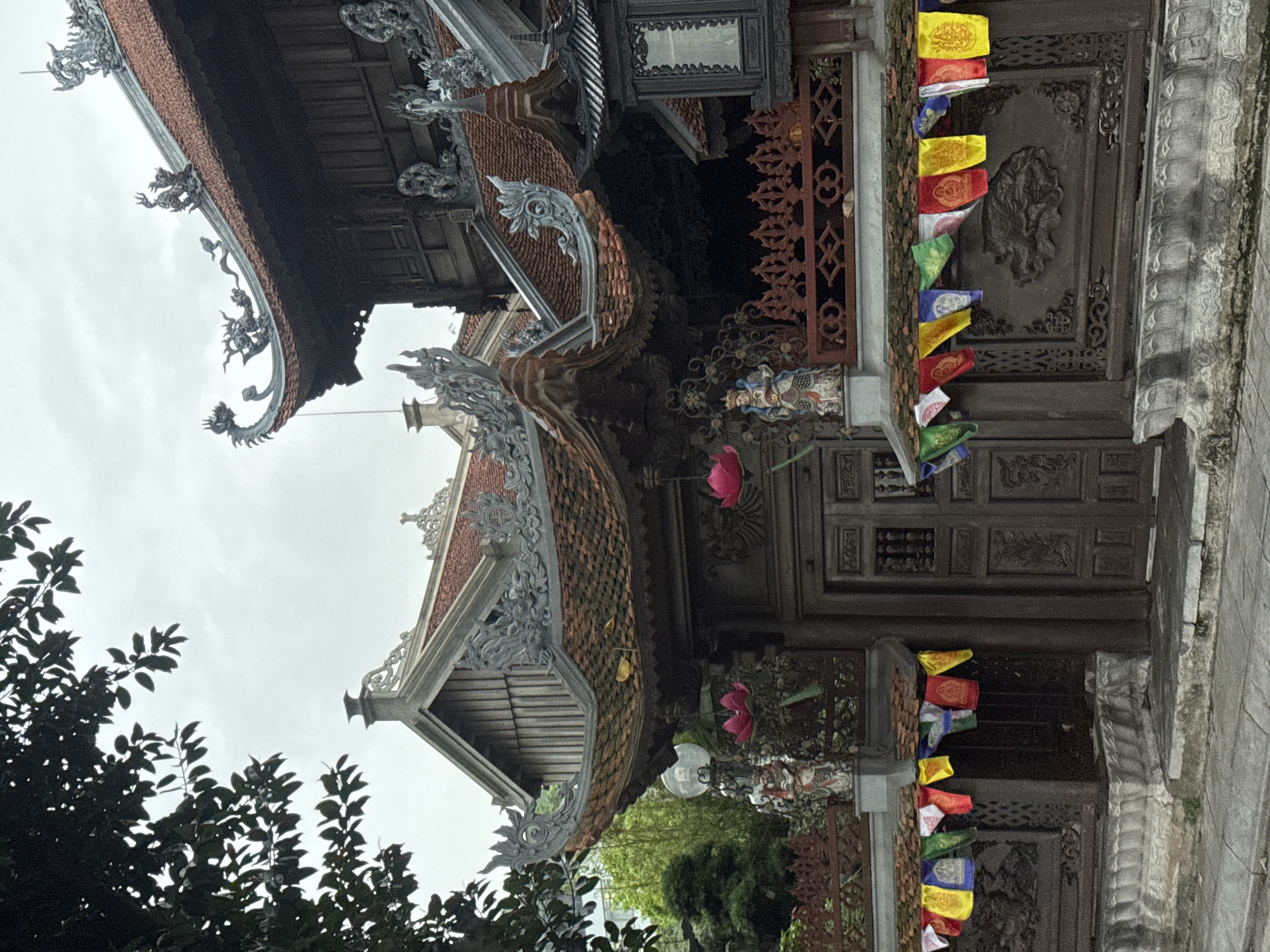 This is a vertical, eye-level, medium shot of the facade of a temple in Hanoi, Vietnam. The building has traditional East Asian architecture, with dark wooden doors, intricately carved walls, and tiered, tiled roofs with ornate dragon decorations. Prayer flags in various colors, including red, yellow, and blue, are strung across the front of the building. A dark green tree branch with leaves is visible in the upper left corner of the image. Two statues of deities in colorful attire stand on either side of the entrance, flanking large, pink lotus flower decorations. The sky is overcast.