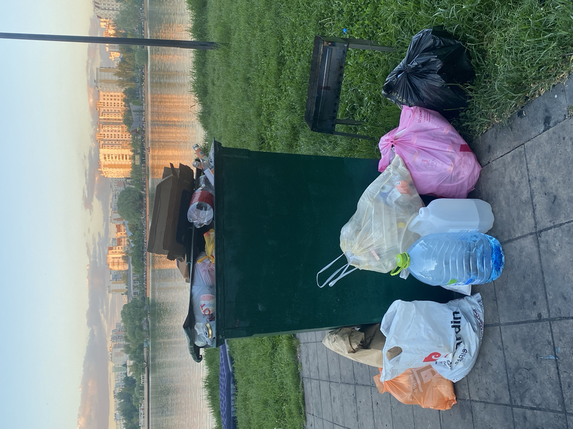 The image depicts a public space in Moscow, Russia, adjacent to a body of water. A green trash receptacle is overflowing with waste. Several bags of trash, including black, pink, and white plastic bags, surround the receptacle. Discarded plastic bottles and other containers are also visible. A grill is placed on the grass near the trash. The surrounding environment includes grass, a paved walkway, and the water with buildings visible on the opposite shore, suggesting an urban or suburban park setting.