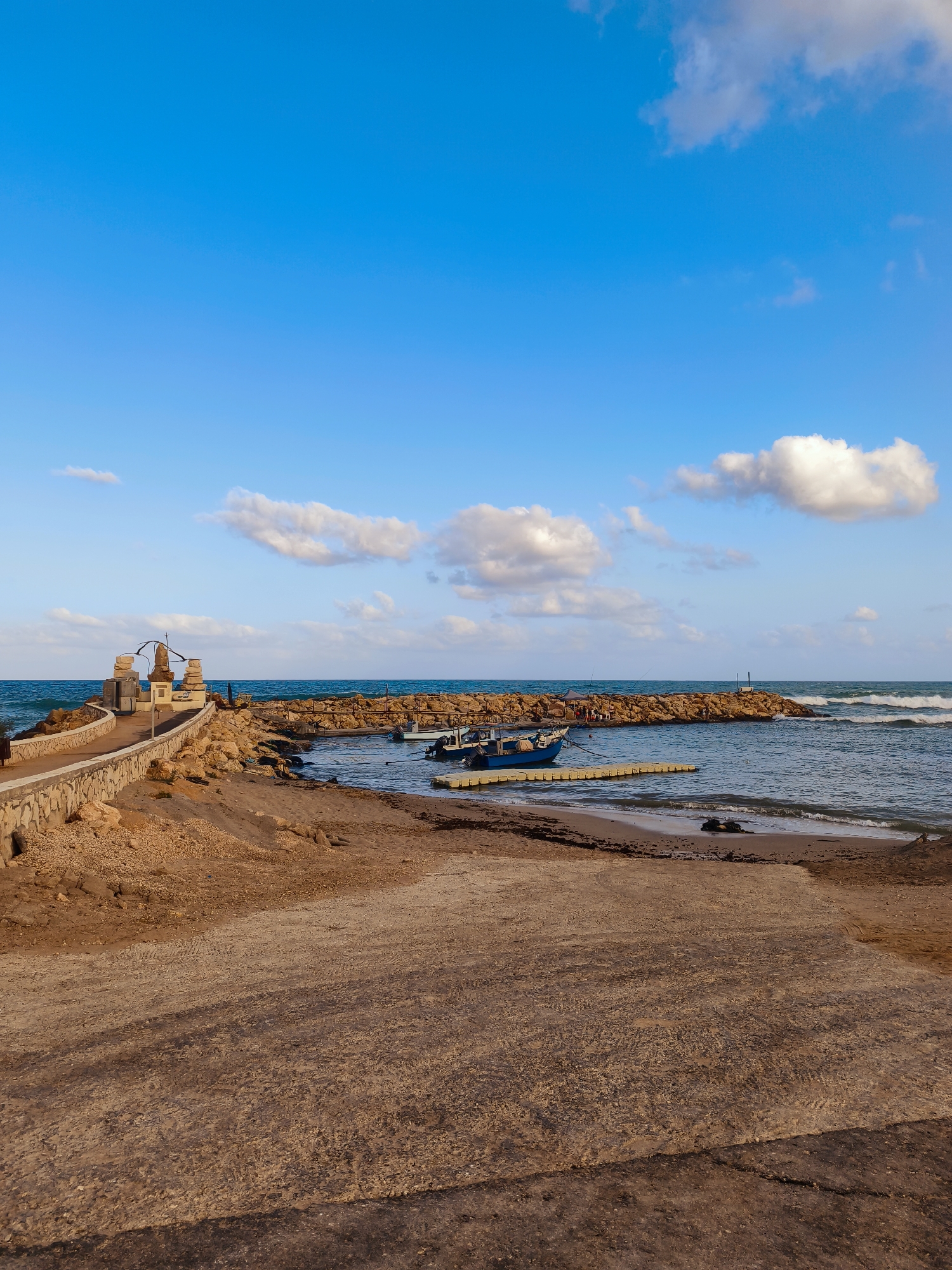 The image depicts a coastal scene in Nahariyya, Israel. In the foreground, a sandy, ramp-like area leads down towards the water. To the left, a stone wall lines a paved walkway that curves around a rocky breakwater. On this breakwater, a monument or sculpture stands, accompanied by several stone structures.
The breakwater itself is constructed of large, rough-hewn rocks and extends into the sea. Within the sheltered cove created by the breakwater, two small boats are visible. One is a blue motorboat tied to a yellow floating dock. A second, smaller white boat is also present. The sea is a deep blue, with moderate waves breaking along the breakwater and washing onto the sandy shore. The sky is a bright blue, dotted with scattered white clouds.
The area appears to be a public space, possibly a small harbor or recreational area. The overall atmosphere is one of calm and natural beauty, with the man-made structures integrated into the coastal landscape. There are no people clearly visible in the immediate foreground or on the beach area.