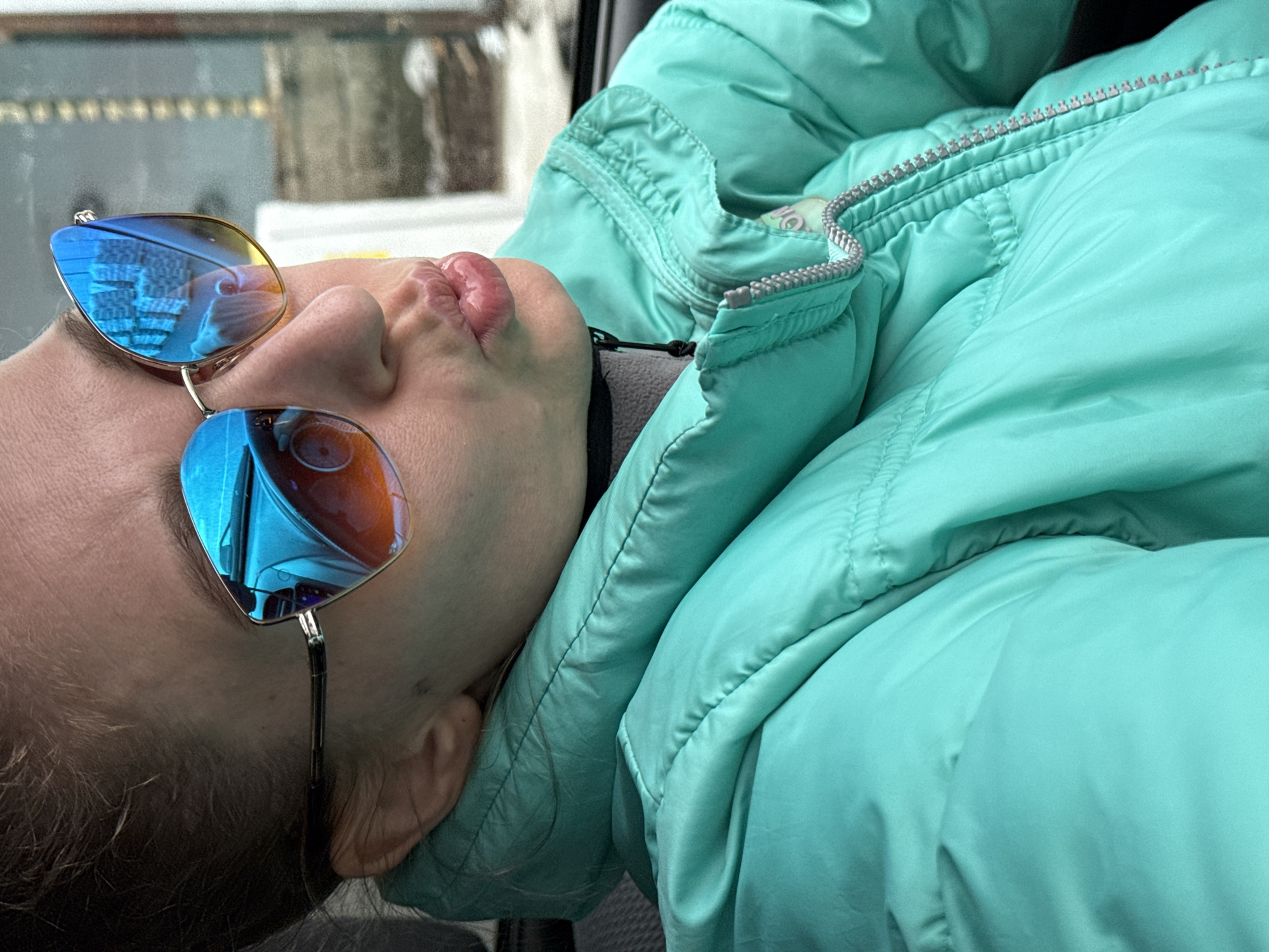 This close-up image captures a person from the shoulders up, leaning back in a relaxed posture. The individual is wearing a vibrant aqua or teal-colored puffer jacket with a silver zipper that runs along the collar. A dark grey or black collar or scarf is visible underneath the jacket. Large, blue-mirrored sunglasses obscure the person's eyes, and their lenses reflect distorted architectural shapes, possibly buildings or structures from an outdoor urban environment. The person's lips are slightly pursed, suggesting a relaxed or playful expression. Light brown hair is visible near the ears and forehead. The background beyond the person is blurred, indicating a shallow depth of field, and appears to be out of focus light and shadows, consistent with being inside a vehicle or resting against a seat. The overall scene conveys a moment of rest or travel, likely in cooler weather, in Chaikovsky, Russia. No clear text is visible in the image.