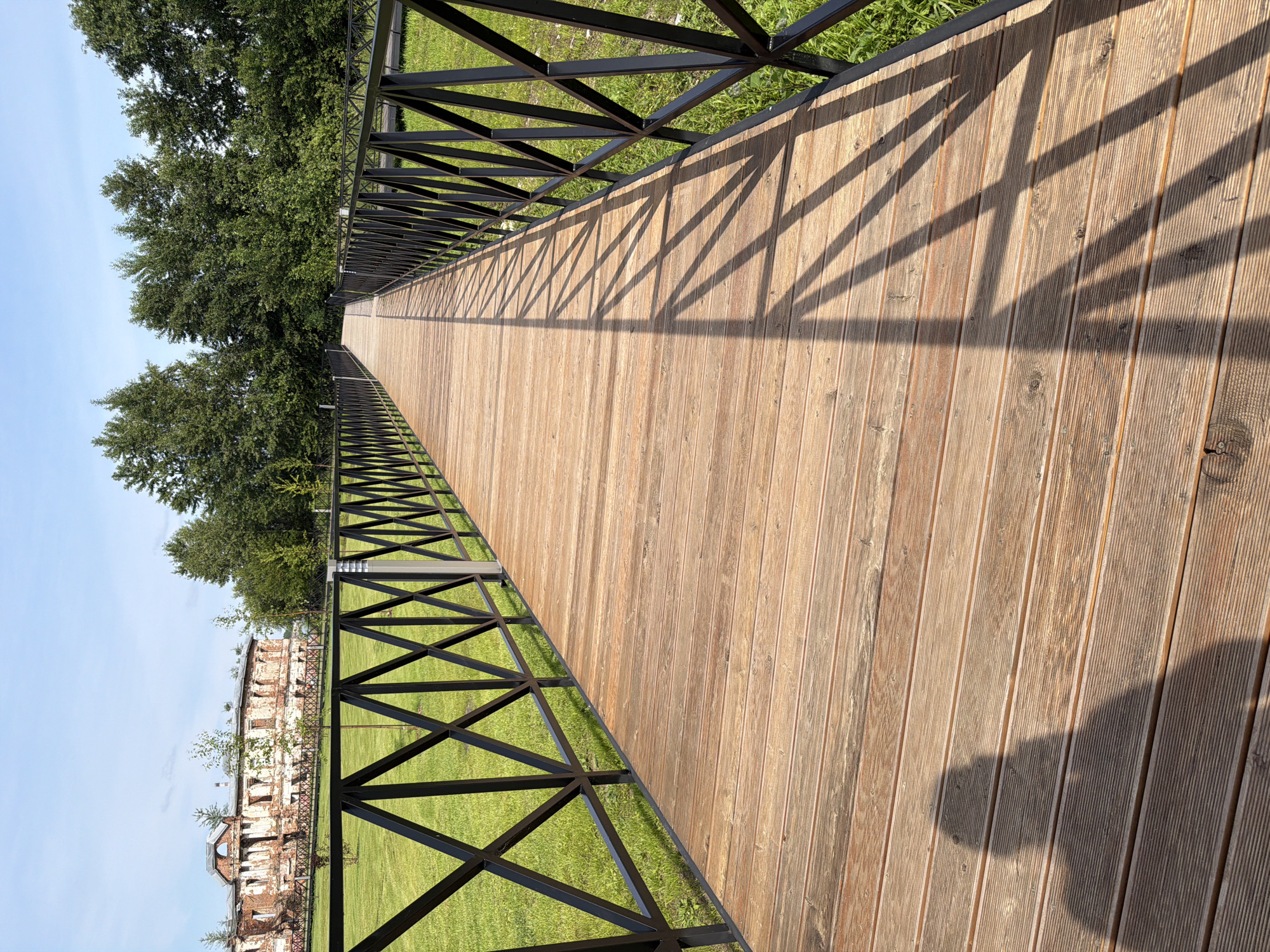 An outdoor, eye-level, medium shot captures a wooden walkway with black metal railings under a clear, bright blue sky. The walkway, made of horizontal wooden planks, recedes into the distance, casting a latticed shadow from the railing onto its surface. To the left of the walkway is a neatly mown green lawn. In the far background, partially visible through trees, stands a large, old, brick building with broken windows and roof, suggesting it is derelict. The building is made of red brick and has a pale, weathered facade. The surrounding environment is characterized by lush green grass and a dense line of deciduous trees bordering the walkway and the building. The shadow of a person is visible on the walkway in the foreground, indicating a human presence. The location is identified as Usolye, Russia.