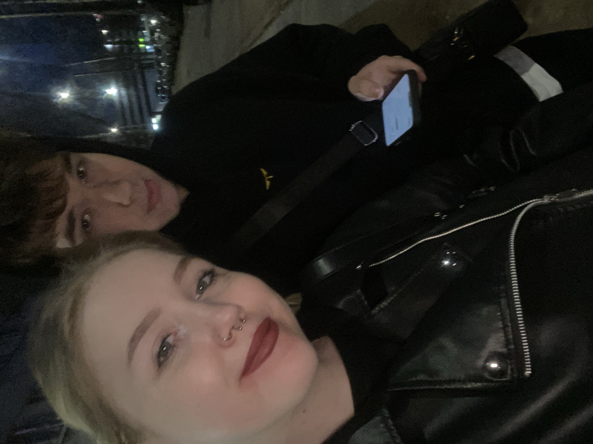 Two people are visible in the image. One person, presumably female, is in the foreground with blonde hair, red lipstick, and a nose piercing. The other person is behind her, partially obscured, wearing a black hoodie and holding a smartphone. They appear to be taking a selfie. The people are wearing black clothes. A black leather jacket is in the foreground, and a black bag with a strap is also visible. The location appears to be urban, with lights and structures in the background. It is night. There is no discernible indication that this is in Izhevsk, Russia.