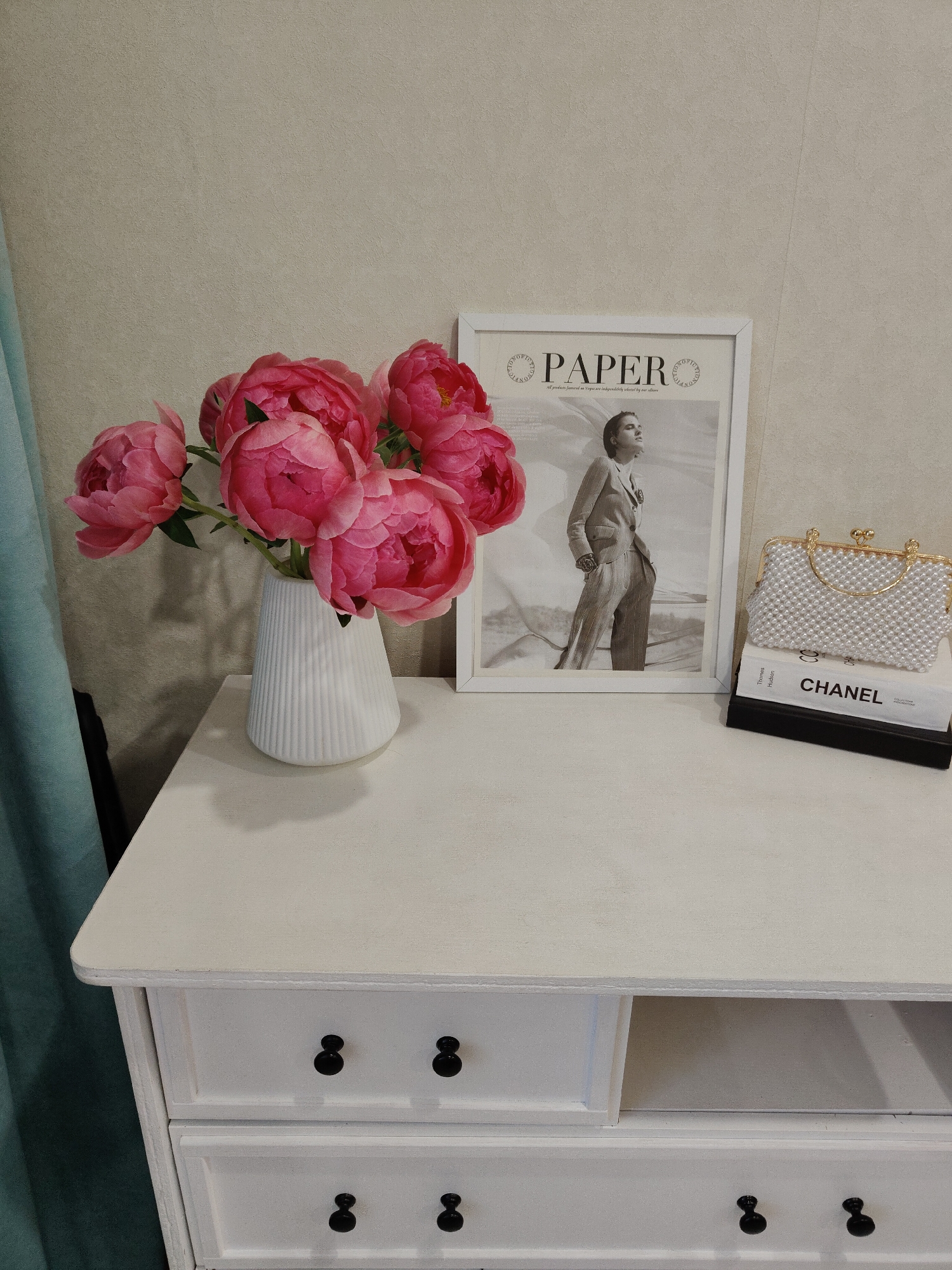 The image displays an indoor scene featuring a white dresser or chest of drawers against a light beige textured wall, in the city of Unknown, Unknown. To the left, a teal curtain is partially visible. The top surface of the dresser holds a decorative arrangement.
On the left, a white, fluted, conical vase contains a bouquet of several vibrant pink peonies with green stems and leaves. To the right of the vase, a white-framed picture stands upright. The picture is a black and white photograph of a woman in a suit, looking upwards, with the word "PAPER" in large capital letters at the top, flanked by circular text.
On the far right, a stack of two books is topped by a white, pearl-beaded clutch bag. The visible book spine reads "CHANEL" with "COLLECTION MODERNE" partially visible below it. The clutch bag has a gold-colored frame and a clasp closure.
The white dresser itself features black, spherical knobs on its drawers. The top right drawer is partially open, revealing a light grey interior. The dresser's surface appears to have a slightly textured or matte finish.