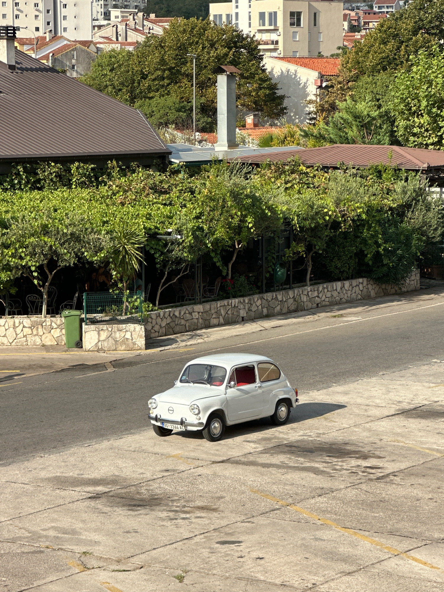 A white vintage car is parked on a concrete lot in Budva, Montenegro. The car is a small, two-door sedan with red interior visible. It has a license plate that reads "BG 3364 AC". The car is casting a shadow to its right. In the background, there is a road, a stone wall, lush green trees, and buildings. One of the buildings appears to be a restaurant with outdoor seating. Further in the background, there are apartment buildings and red-tiled roofs characteristic of the region. The sky is clear and the sun is shining, casting distinct shadows. The scene appears to be set in a semi-urban environment, likely during the daytime.