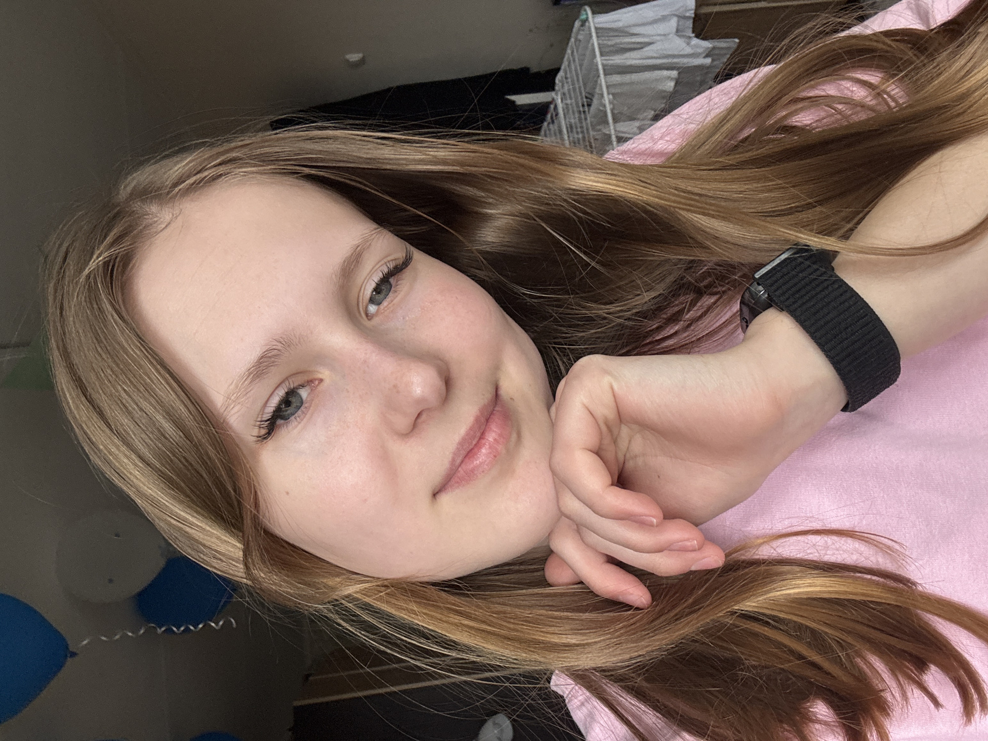 A medium close-up portrait shows a young woman with long, light brown hair with blonde highlights, lying down and looking directly at the camera with a gentle, soft smile. Her head is tilted slightly, resting on her right hand, with her fingers lightly curled against her cheek. She has clear skin with faint freckles visible on her nose and cheeks, light-colored eyes (appearing blue-green), and defined eyelashes. Her lips are a natural light pink.
She is wearing a pink top or lying on a pink fabric surface, and a black watch with a woven fabric strap is visible on her left wrist. Her long hair is spread out around her head and shoulders. The background elements suggest an indoor, domestic setting, possibly a home in Chaikovsky, Russia. To the upper right, a white clothes drying rack is partially visible. To the lower left, two blue balloons with a curly white string are reflected in a dark, glossy surface, possibly a floor or furniture. The lighting is soft and even, suggesting an indoor environment. Her relaxed pose and mild smile convey a calm and pleasant emotion. There is no visible text in the image.