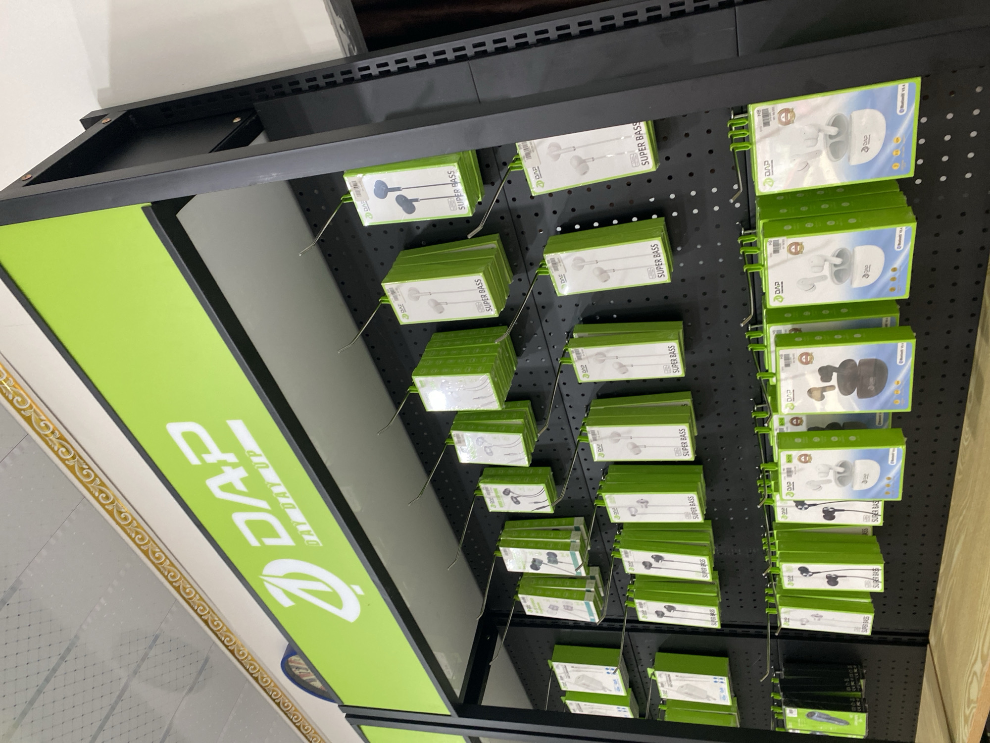 The image shows a retail display of electronics, specifically earphones and earbuds, in a store. The products are packaged in green and white boxes and are hung on metal hooks attached to a black pegboard wall. The store's branding, "DAP DAY DAY UP," is visible on a lime green sign at the top of the display. The products are neatly arranged in rows, with different types of earphones and earbuds visible. The overall impression is of a well-organized retail environment. The location of Ta', Indonesia, is provided as context.