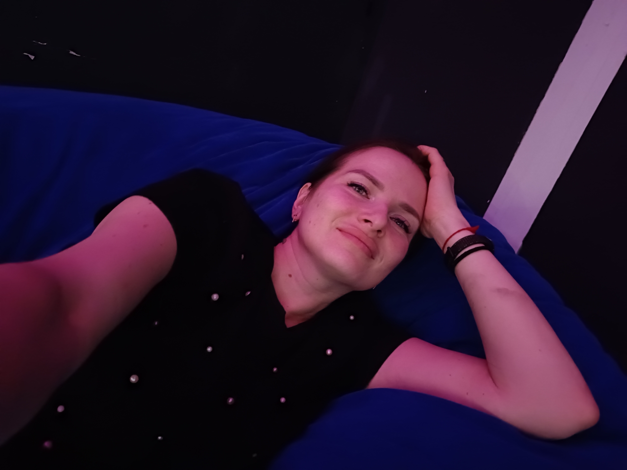A young woman is captured in a selfie, relaxing indoors on a large blue bean bag chair. She lies on her side, resting her head slightly with her left hand near her ear, and her right arm bent. She wears a black shirt adorned with small, light-colored embellishments, possibly beads or pearls. A black wristband or watch with a red string tied around it is visible on her right wrist.
The woman gazes directly at the camera with a gentle, contented smile, suggesting a relaxed and pleasant mood. Her skin is bathed in a soft, artificial pink and purple glow, indicating the use of colored lighting in the room. This lighting creates a dim, cozy, and somewhat atmospheric environment, suggesting the picture was taken in the evening or at night. The background is predominantly dark, with a distinctive diagonal strip of white or light purple on the right, possibly a wall feature or ceiling element. The overall setting is informal and comfortable, likely a lounge area or a personal space. The scene is located in Chaikovsky, Russia.