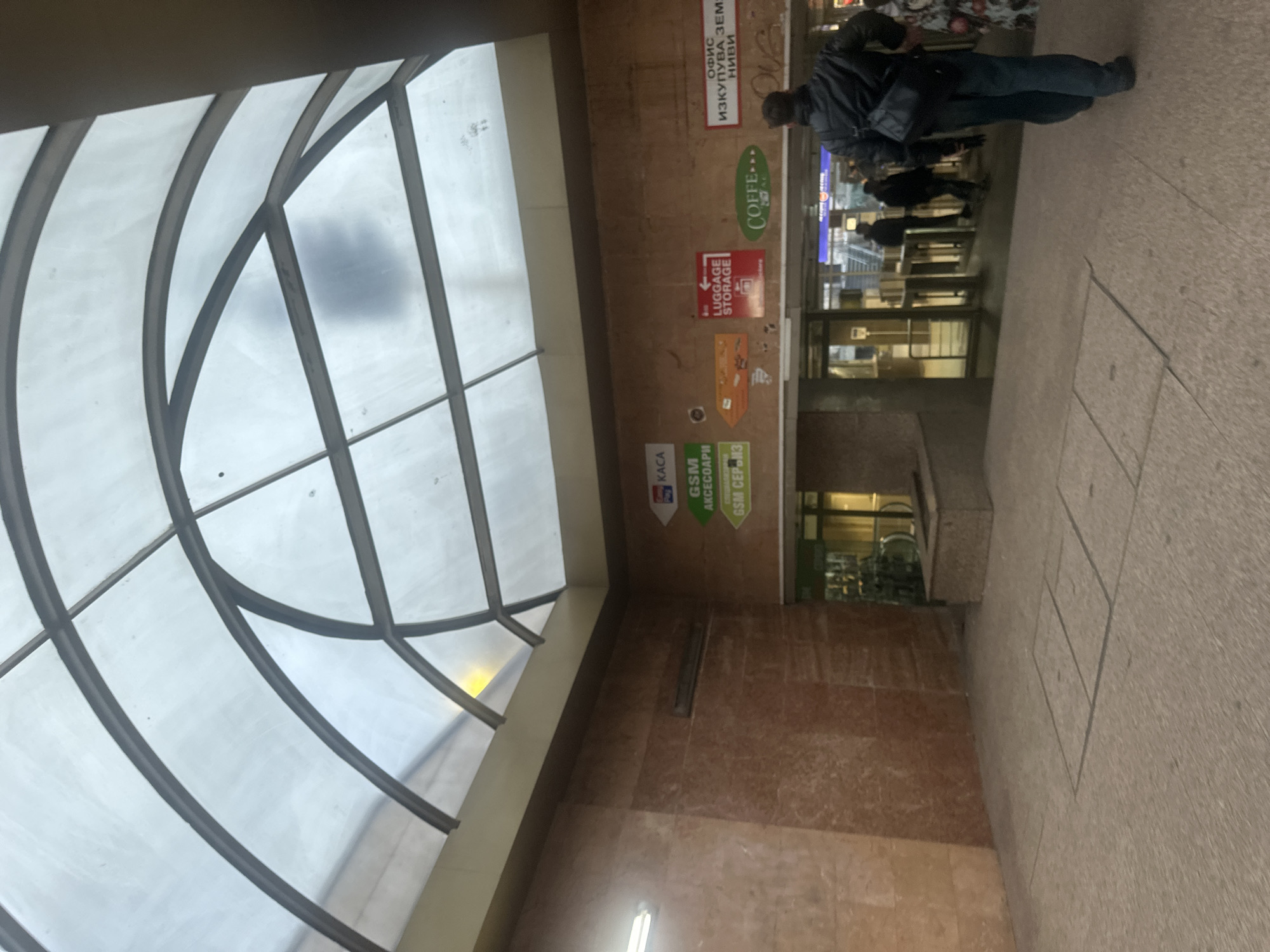 The image shows the entrance to a subway station in Sofia, Bulgaria. The scene is captured from an indoor perspective. The ceiling has a curved, transparent design with a metal frame. The walls are tiled in brown and are covered with various advertisements and informational signs in Bulgarian. People are walking through the entrance and exit, most notably a man in a dark jacket. The floor is paved with large concrete tiles.