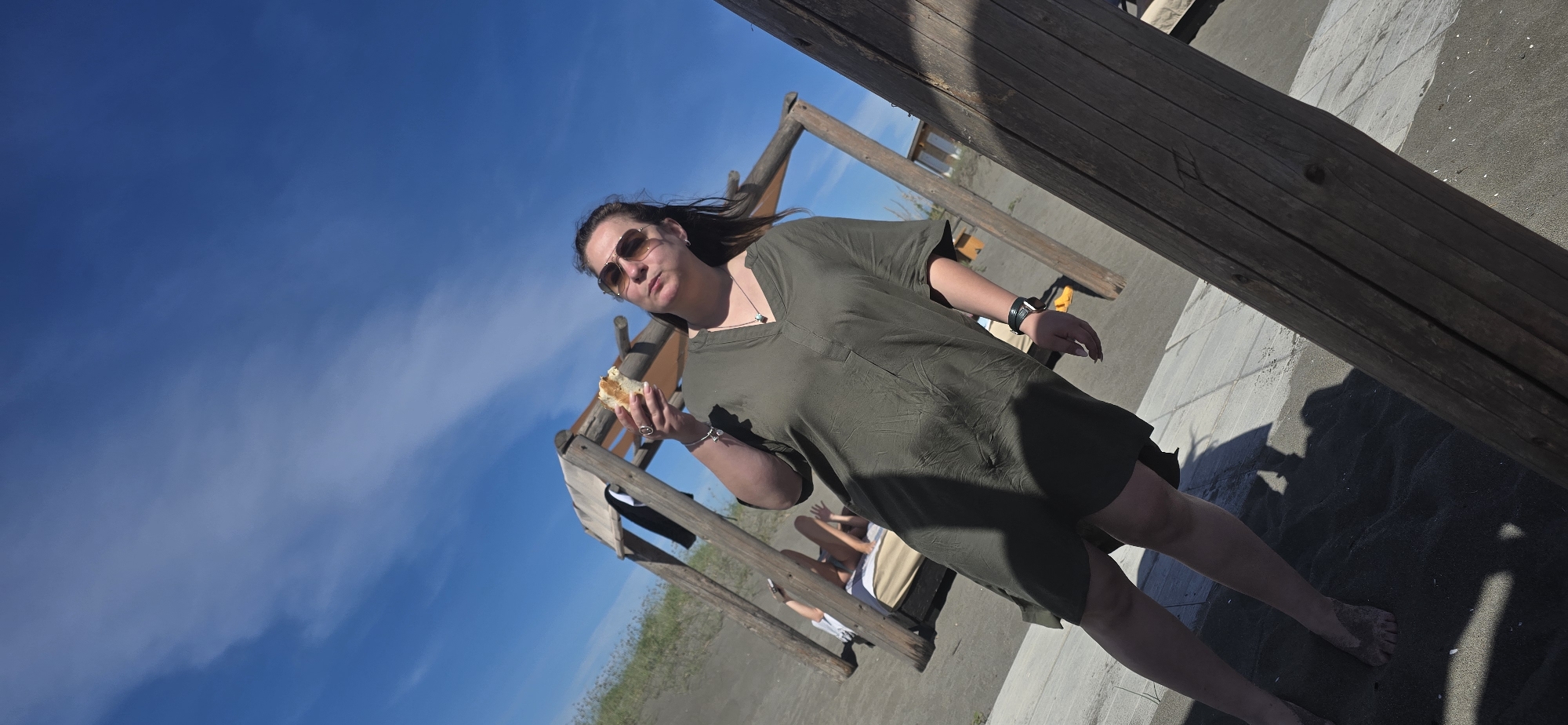 A woman stands on a beach in Gornji Štoj - Shtoji i Epërm, Montenegro, holding a sandwich. She wears sunglasses and a dark green dress. Behind her is a wooden structure with people sitting in it. The sky is blue with wispy clouds. A large wooden beam is in the foreground, partially obscuring the view. The ground is a mix of sand and concrete.