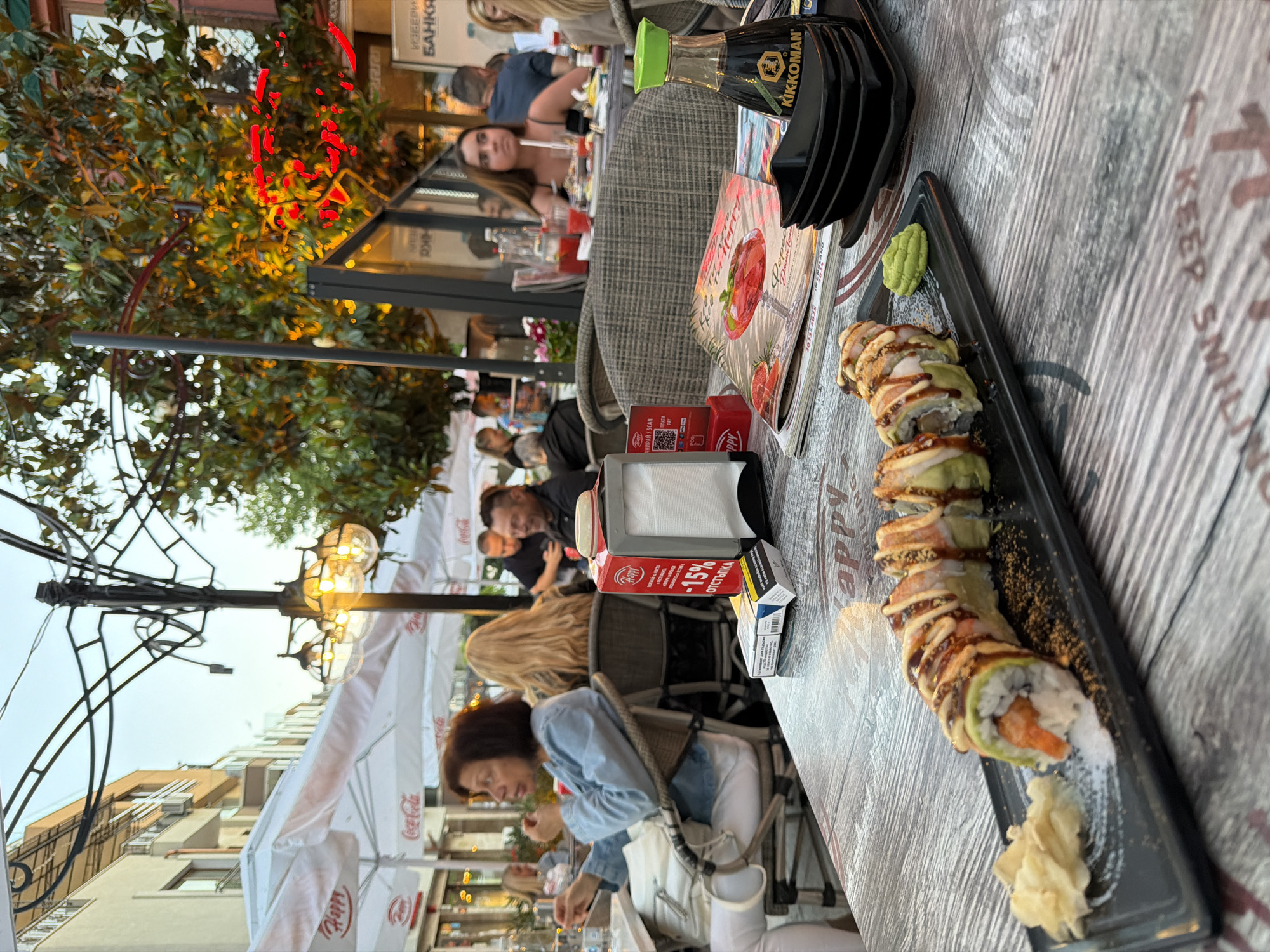 The image depicts an outdoor seating area of a restaurant, likely located on a street in Burgas, Bulgaria. The time appears to be late afternoon or early evening, given the soft lighting and the illuminated streetlights and tree lights. A long sushi roll is prominently displayed on a black plate in the foreground, accompanied by pickled ginger and wasabi. In the background, several patrons are seated at tables. A woman in a denim jacket is seated at the table in the foreground, facing left. Another woman with blonde hair is seated behind her. Further back, other diners are visible, some in conversation. The restaurant's outdoor setup includes white umbrellas, tables, and chairs. A bottle of Kikkoman soy sauce is also on the table near the sushi. The overall atmosphere suggests a casual dining experience.