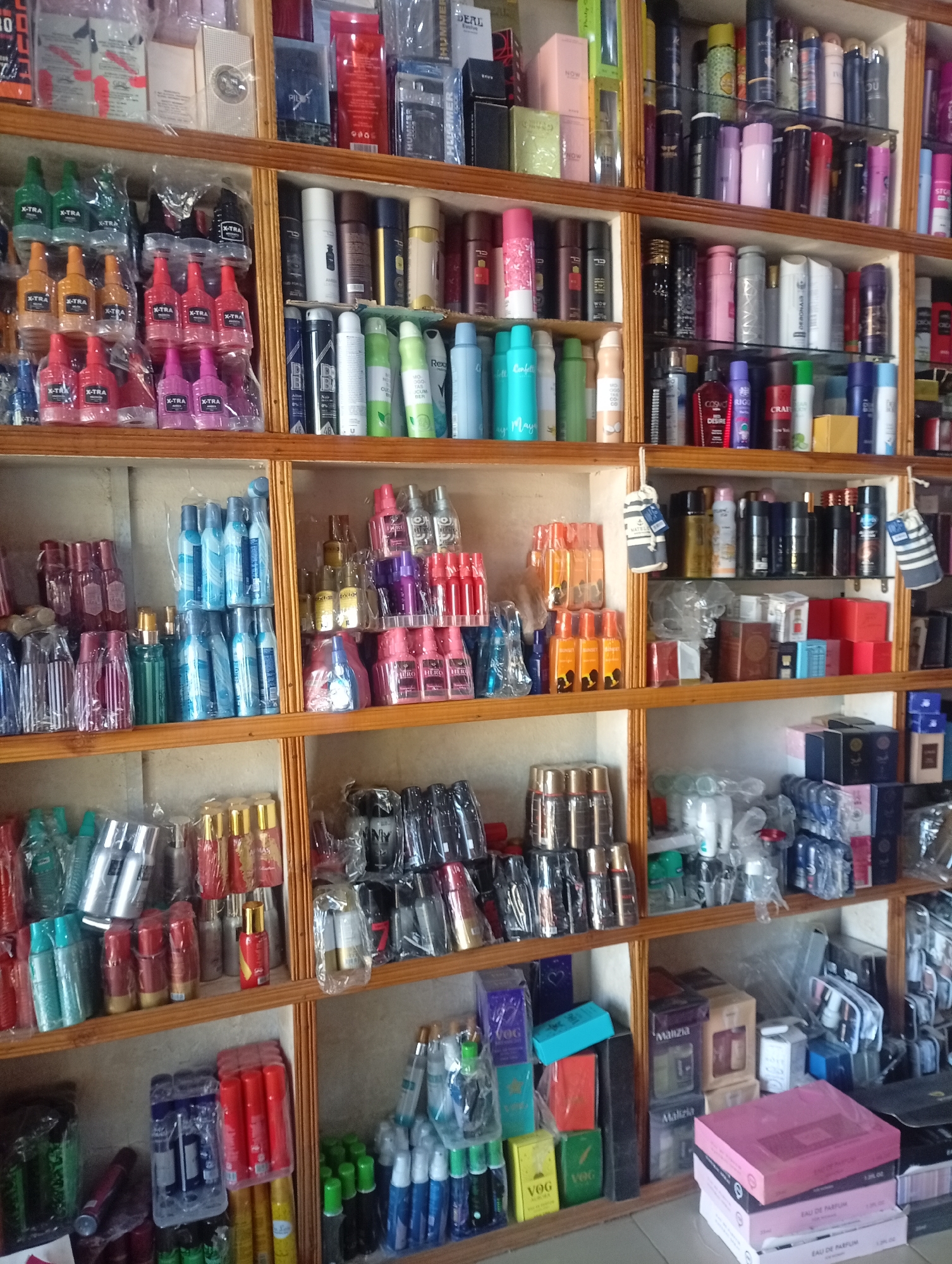 This is a detailed photograph of a store shelf in Bukuru, Nigeria, displaying a wide assortment of perfumes and personal care products. The shelves are made of light-colored wood and are filled with various bottles and boxes of perfumes, colognes, and deodorants. The products are arranged in rows, with some items still in their original packaging. The lighting suggests an indoor setting, likely a retail environment. The image captures the abundance of products available for sale, showcasing a variety of brands and sizes.