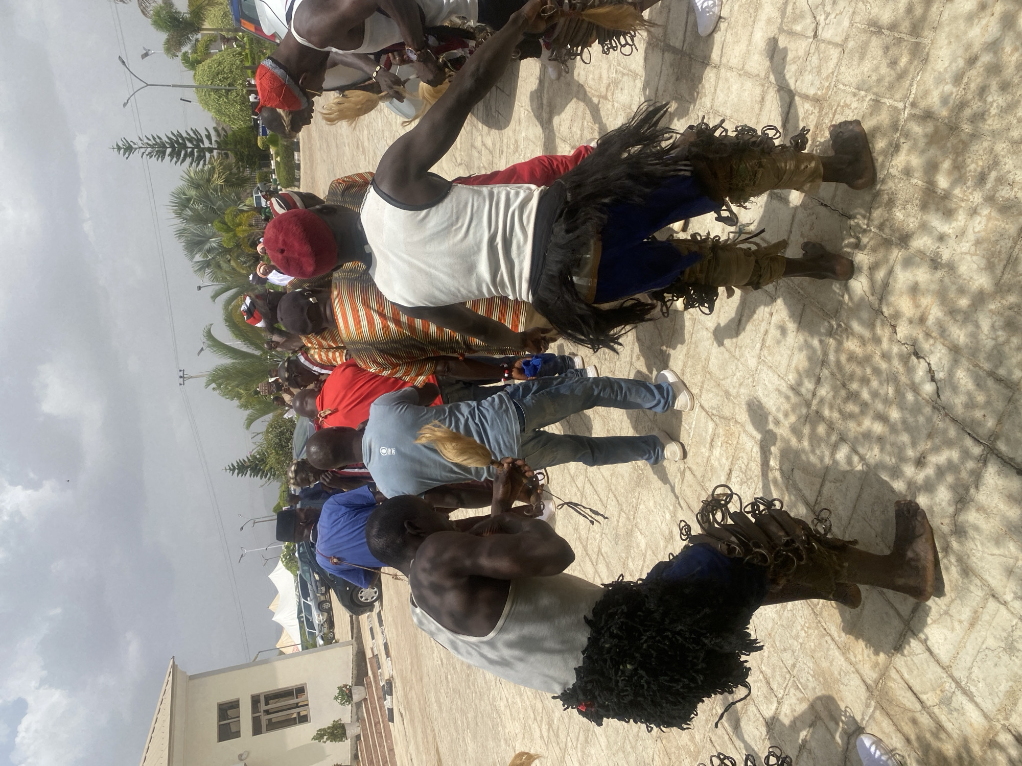 A cultural event is underway in Jos, Nigeria, featuring a gathering of people on a paved open area. A group of men are engaged in a traditional performance, dressed in attire including white tank tops, blue pants, and distinctive leg coverings adorned with dark, shaggy fringe and looped embellishments. Some wear traditional hats, including red caps and striped headgear. The performers hold objects resembling blonde tassels.
The environment includes a building, trees, and light poles. Automobiles are parked in the background near a white tent. The sky is overcast.