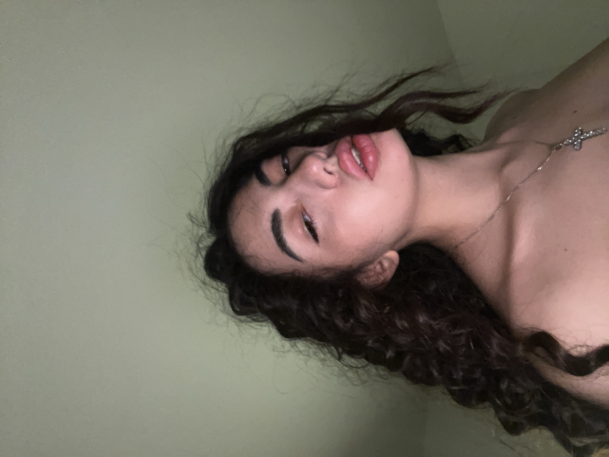 The image is a close-up, low-angle selfie of a young woman with dark, curly hair. She is looking directly at the camera with her lips slightly parted, revealing her teeth. She is wearing a silver cross necklace. The background is a plain, pale green wall. There is no indication of the location being Chotěšov, Czechia.