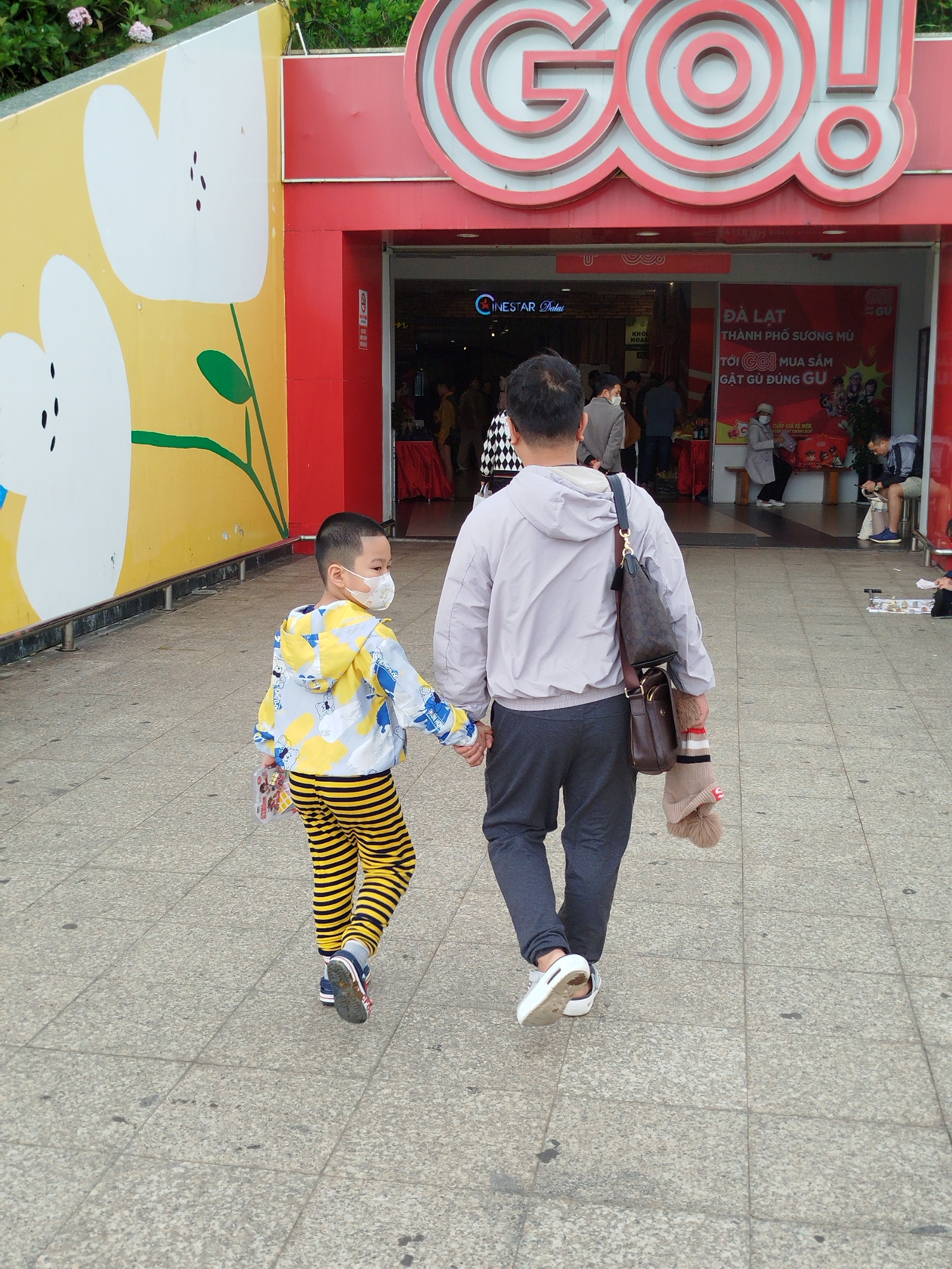 A man and a young boy are walking hand-in-hand away from the camera on a paved walkway outside a building with large red and white "GO!" signage. The boy, on the left, wears a yellow and white hooded jacket with a cartoon pattern, and black and yellow striped pants. He is wearing a white face mask and holding a small item in his left hand. The man, on the right, wears a light gray hooded jacket and dark gray pants. He has a brown messenger bag over his left shoulder and is carrying a tan knit hat with a fluffy pom-pom in his right hand. The building entrance behind them shows several people inside, and a red poster with Vietnamese text. The walkway is made of gray square tiles. To the left of the walkway is a yellow wall decorated with large white floral patterns. This scene is consistent with an entrance area of a commercial establishment, possibly a shopping center or entertainment venue, in Da Lat, Vietnam, given the signage and building style often seen in such tourist-oriented cities.