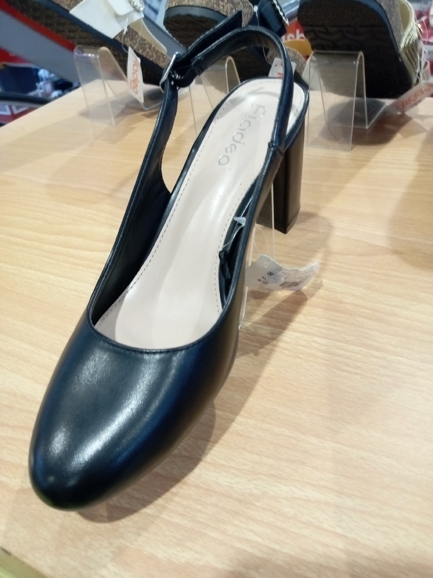 A single black slingback high-heeled shoe is displayed on a light brown wooden surface. The shoe features a pointed toe and a thick, block heel. A white tag with the brand name "Fladeo" is visible on the inner sole, and another price tag hangs from the strap. In the background, out of focus, other footwear is displayed on clear plastic stands. The scene appears to be an indoor retail environment, likely a shoe store. This image was taken in Batam, Indonesia.