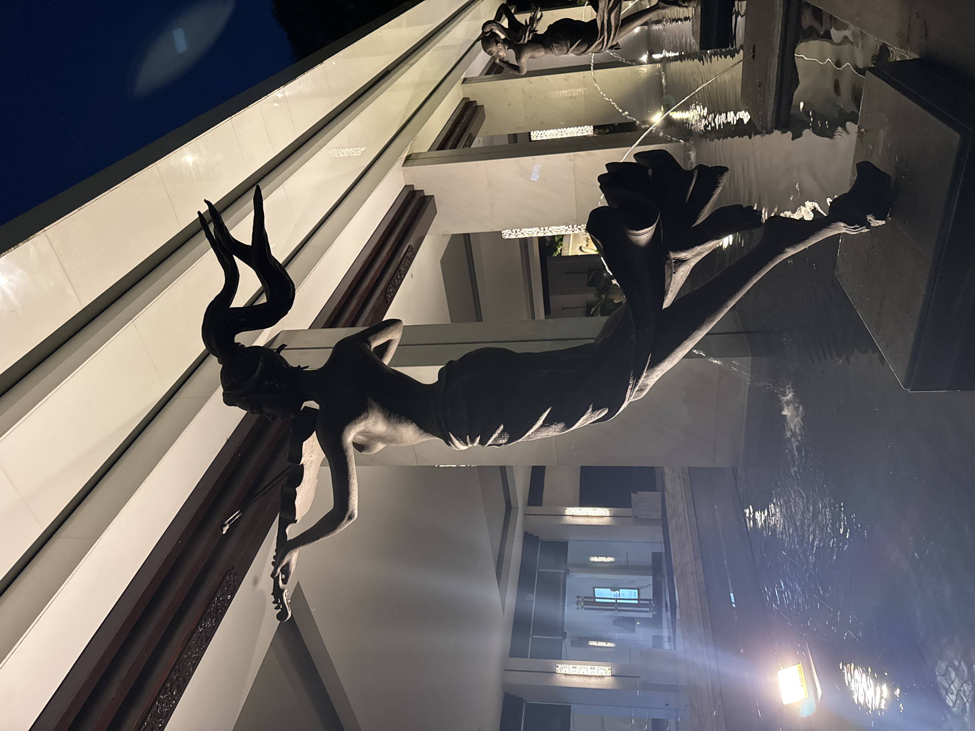 This media file captures a captivating night scene featuring a multi-level modern building adorned with an elaborate water feature and striking sculptures. The image is taken from a low, upward-looking perspective, emphasizing the height of the architecture and the dramatic interplay of elements.
The central subjects are several dark, stylized sculptures of human-like figures, predominantly women. The most prominent sculpture in the foreground, positioned as if floating or gracefully moving, has distinct antler-like structures emerging from its head, giving it a mythical or nature-inspired appearance. Other figures are seen further back, integrated into the water element, one appearing to dive into or emerge from the water with outstretched arms. The sculptures have a dark, possibly bronze or stone-like finish, and are often silhouetted against the illuminated architecture or partially lit by artificial light.
The building itself showcases a clean, modern aesthetic with smooth, white or light-colored surfaces forming distinct horizontal levels or balconies. These levels are punctuated by dark, ornate panels or structures, possibly made of wood or metal, adding a touch of traditional or intricate detail. The building's reflective surfaces and the dark sky suggest it is nighttime.
A significant part of the scene is the body of water, likely a pool or large fountain, situated at various levels and around which the sculptures are placed. Water can be seen cascading or flowing, creating ripples and reflections of the building's lights, which cast a bright glow and sharp contrasts across the scene. The artificial lighting, including strip lights and spotlights, illuminates the architectural details and highlights the textures of the sculptures and the movement of the water.
The overall atmosphere is serene and artistic, presenting a grand architectural and sculptural installation. No legible text is visible in the image. Given the location context of Kuta Selatan, Indonesia, this likely depicts a sophisticated resort or public art installation.