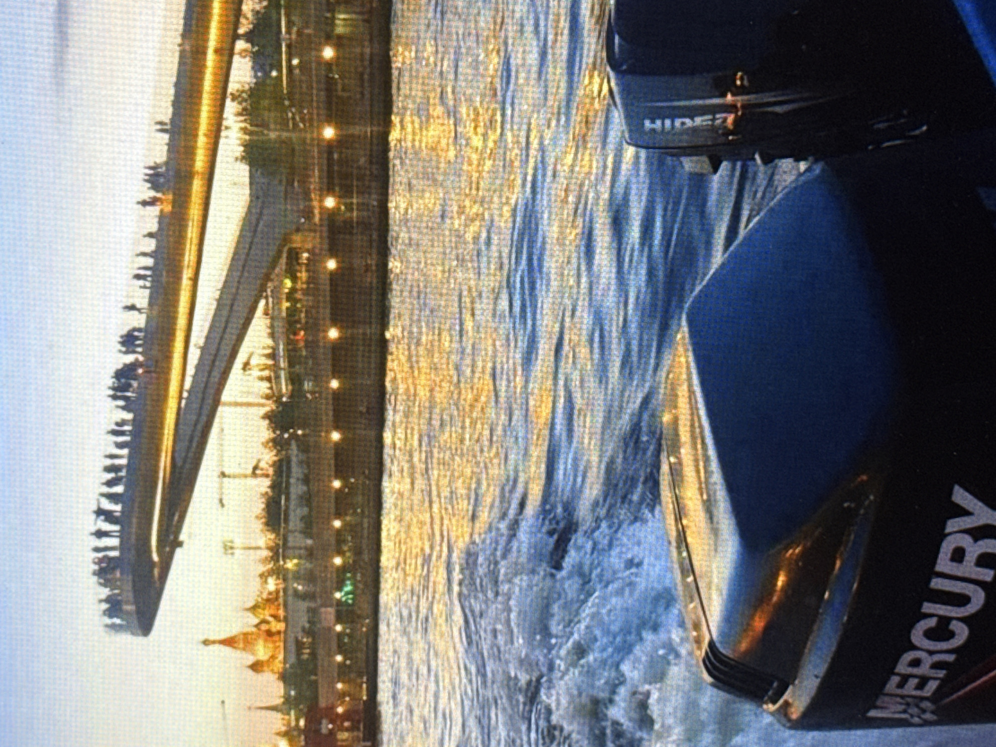 The image depicts a sunset scene over a body of water, likely a river, with a modern pedestrian bridge in the background. The bridge, illuminated by warm lights, is crowded with people silhouetted against the sky. In the distance, prominent golden-domed buildings, possibly churches or cathedrals, are visible. The foreground shows a portion of a boat, with its engine cover prominently displaying the "MERCURY" logo. The water is choppy, creating white wakes, indicating the boat is in motion. The sky transitions from a light blue at the top to warmer orange and yellow hues near the horizon, reflecting on the water.