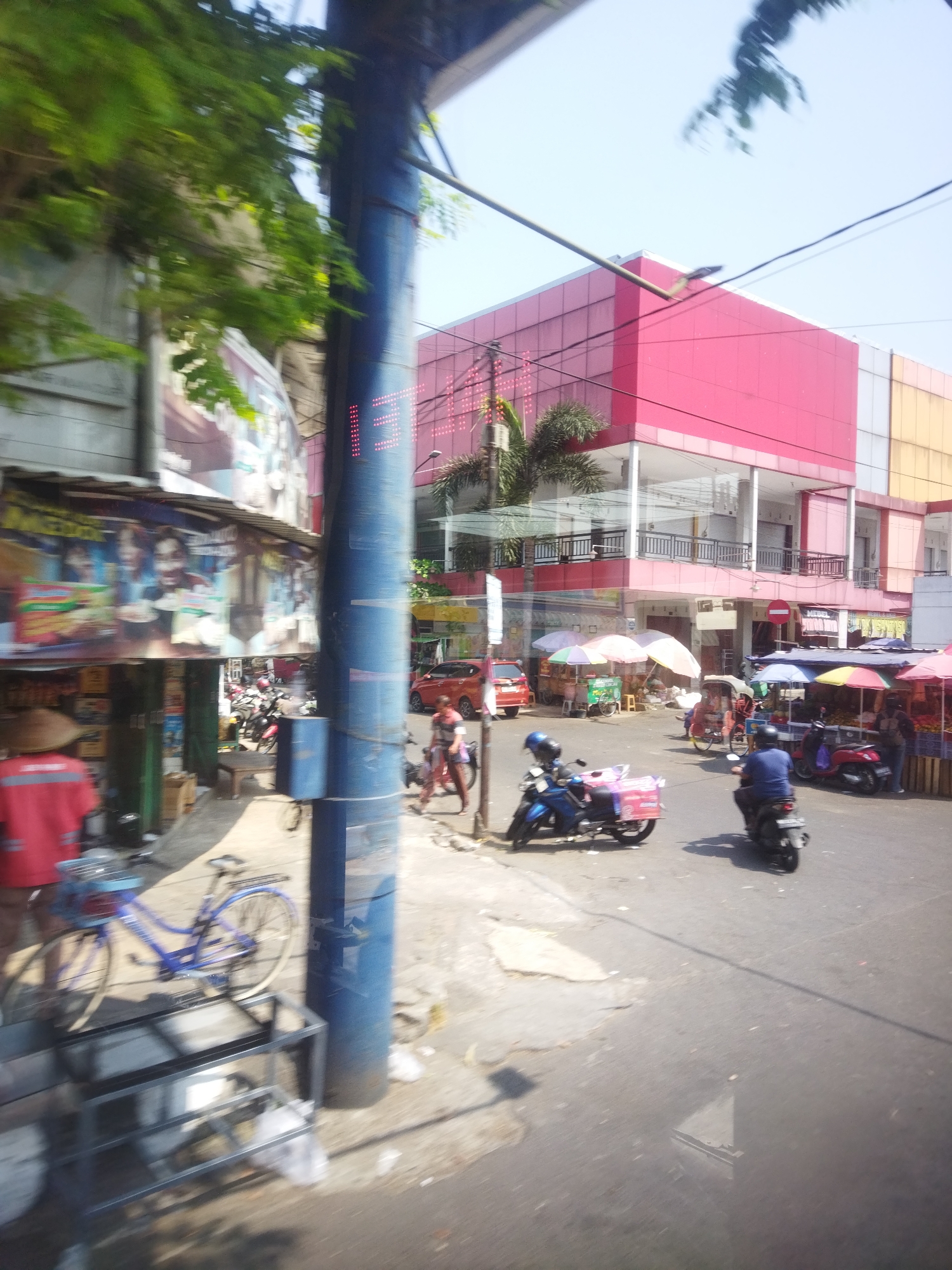 The image captures a street scene in Sukoharjo, Indonesia, likely a commercial area. In the foreground, a blue bicycle is parked next to a large blue utility pole. To the left, a person wearing a red shirt and a conical hat is partially visible, and further back, there are several stalls with colorful awnings selling food or goods, with posters advertising products. The background features a prominent building with a bright pink facade and a lighter colored section above it, suggesting it might be a mall or large shop. Several motorcycles are present on the street, with some people riding them and others parked. One motorcycle in the foreground is blue, and another further back is red. The street appears to be paved, with some debris on the ground. The overall impression is a bustling urban environment on a sunny day, with a mix of traditional elements like the conical hat and modern architecture.