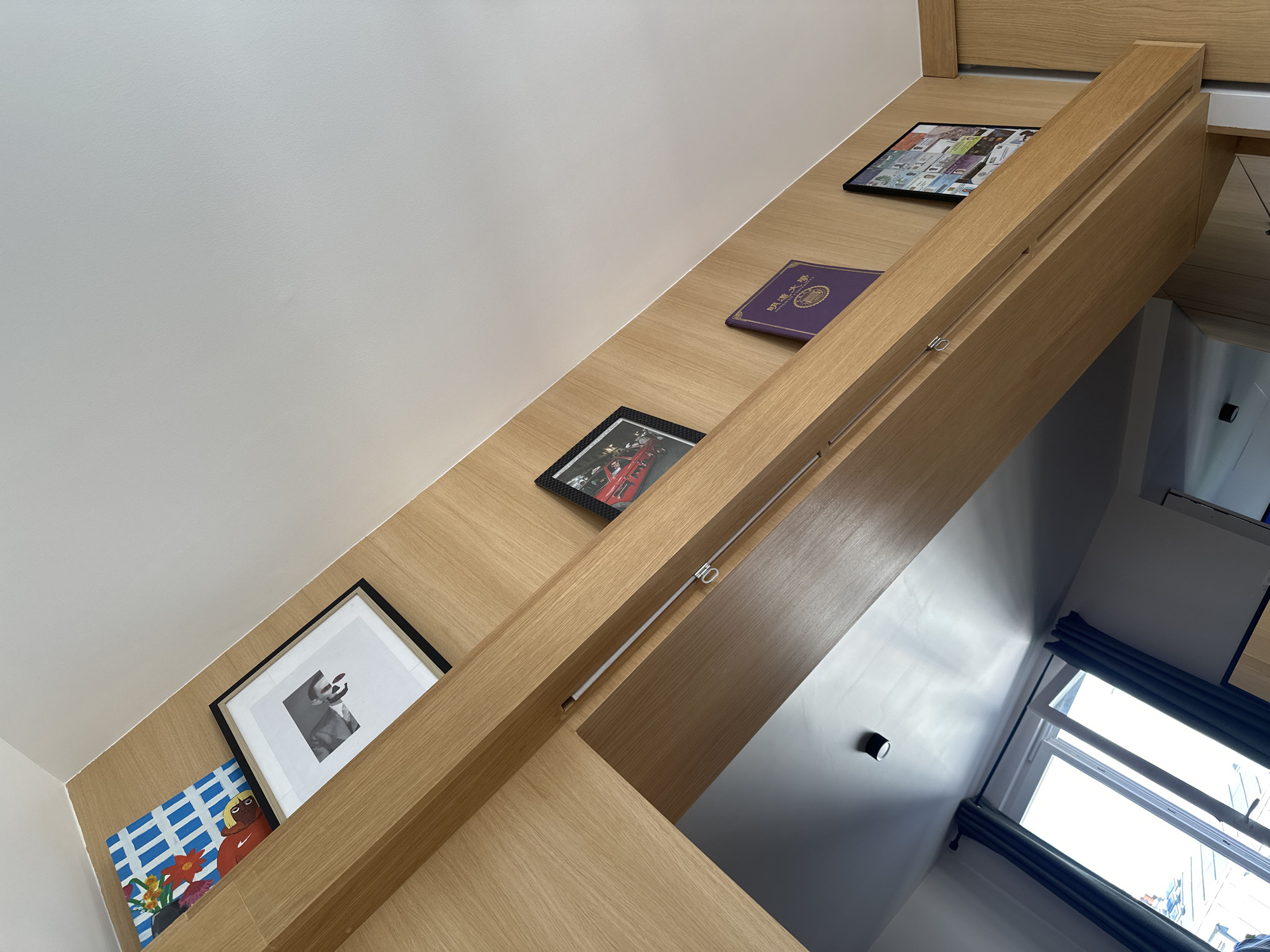 A low-angle interior shot shows a section of built-in wooden shelving against a white wall. The shelves are adorned with various framed pictures, a book, and an artwork. The lighting suggests daylight entering from a window partially visible in the lower right corner. The shelves are constructed of a light-colored wood. The perspective is from below, looking up at the shelves which are mounted along the wall and angling toward an opening to another room, where a window with blue curtains is visible. Saint-Ouen-sur-Seine, France is not directly discernible from the image content.