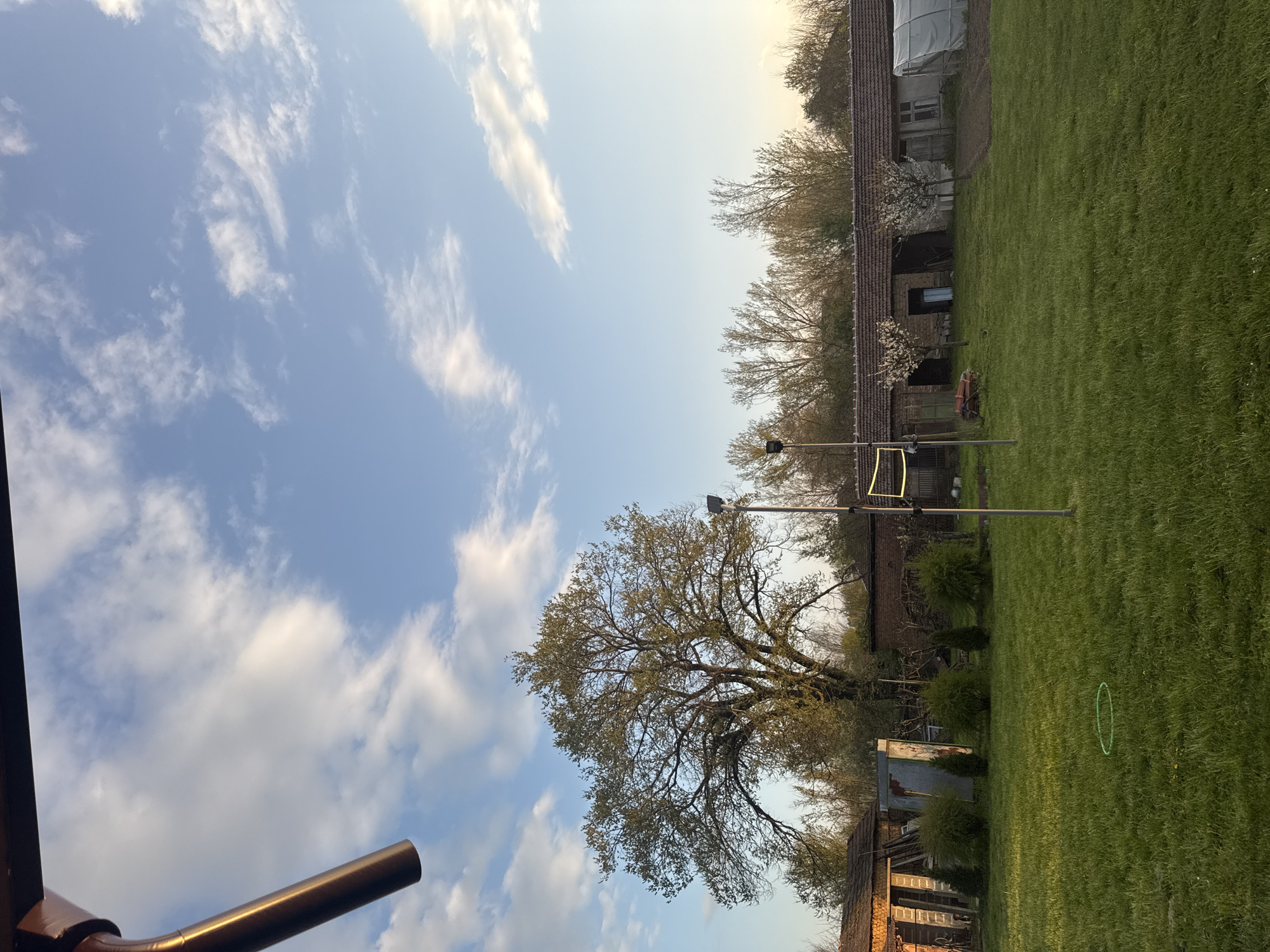 The image shows a rural landscape in Lehchevo, Bulgaria. A large tree dominates the mid-ground, partially obscuring old brick buildings with tiled roofs. A green lawn stretches across the foreground and toward the buildings. A volleyball net is erected in the lawn, its poles visible against the backdrop of a building. The sky is light blue with scattered white clouds. A brown gutter is in the lower left corner of the frame.