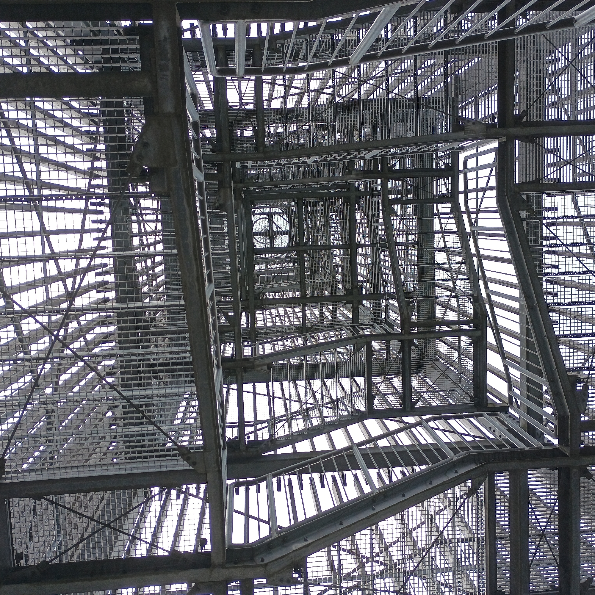 This is a low-angle, wide shot of the interior of a large metal observation tower, likely the Möhnesee Water Tower, located in Möhnesee, Germany. The image is dominated by a complex network of interconnected steel beams, stairs, and platforms, all constructed with a grid-like metal grating. The perspective looks upwards, revealing multiple levels of the tower's structure receding into the bright, overcast sky. There are no people visible in the image. The metalwork is weathered and shows signs of industrial construction. The scene is stark and geometric, emphasizing the engineering and scale of the structure.