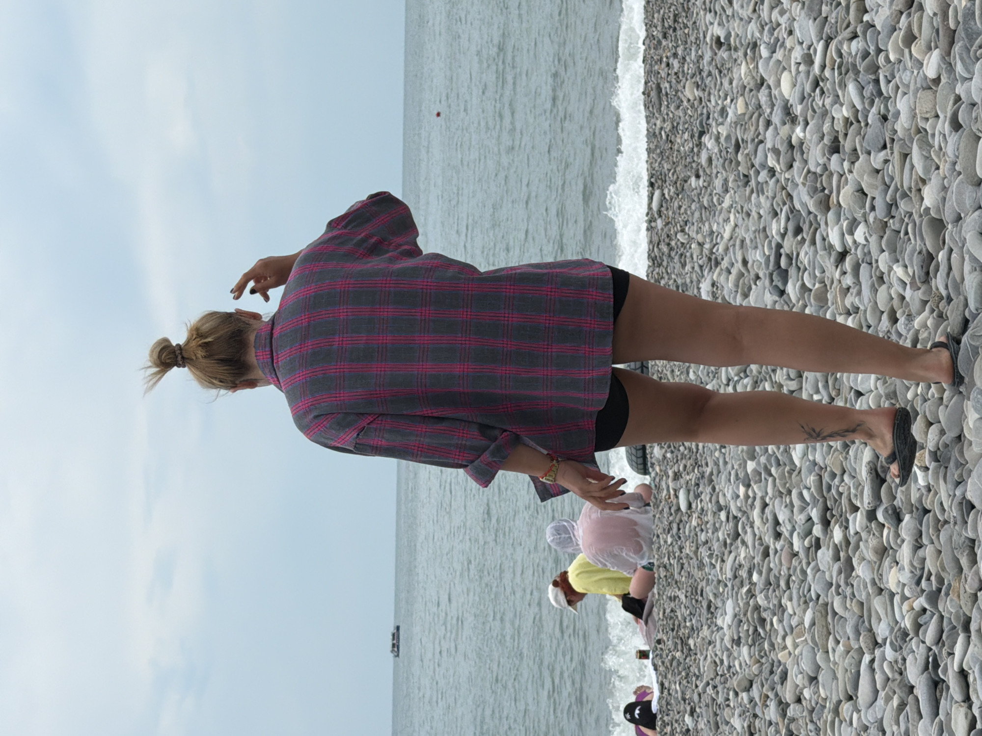 The image depicts a pebble beach in Sochi, Russia, with a woman standing in the foreground, facing away from the camera. She has her hair tied up in a bun and is wearing a pink and grey plaid shirt and black shorts. Her right hand is raised, as if gesturing. In the background, several other people are seated on the beach near the water, some appearing to be wet. The sea is calm with small waves. A boat is visible in the distance on the water. The sky is overcast.