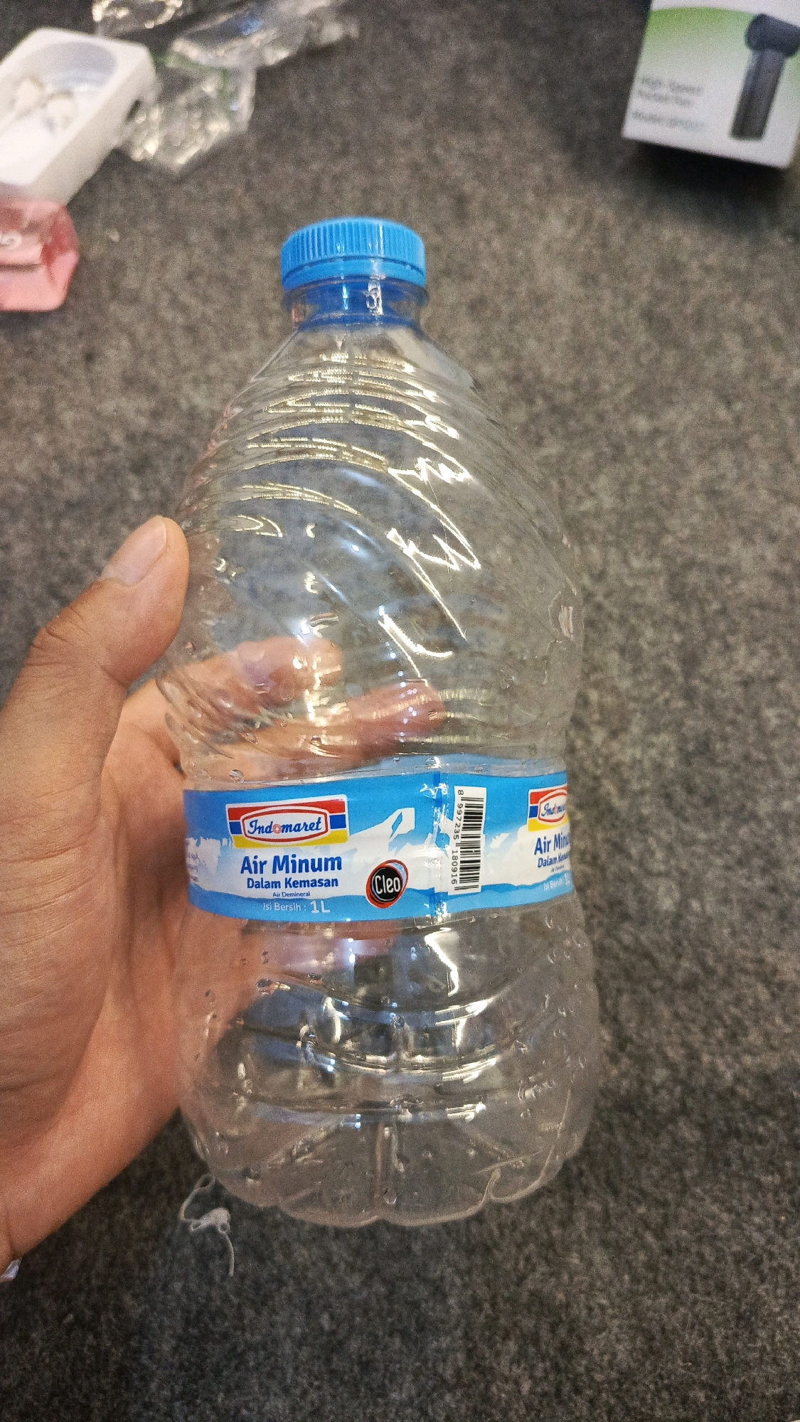 A clear plastic bottle with a blue cap, containing water, is held by a person's left hand. The bottle has a blue and white label with text in Indonesian. The label reads "Indomaret Air Minum Dalam Kemasan" which translates to "Indomaret Packaged Drinking Water". Below this, it states "Air Demineral" (Demineralized Water) and "Isi Bersih: 1L" (Net Content: 1L). The background appears to be a textured gray surface, possibly carpet or a similar material. The scene is indoors, with some out-of-focus objects in the background, including a white plastic container and a cardboard box with an image of a black object on it. The overall lighting suggests an indoor environment. The presence of the Indomaret branding suggests the location could be in Indonesia, and specifically if this were in Babadan, Indonesia, it might be near an Indomaret convenience store or in a context where such products are commonly found.