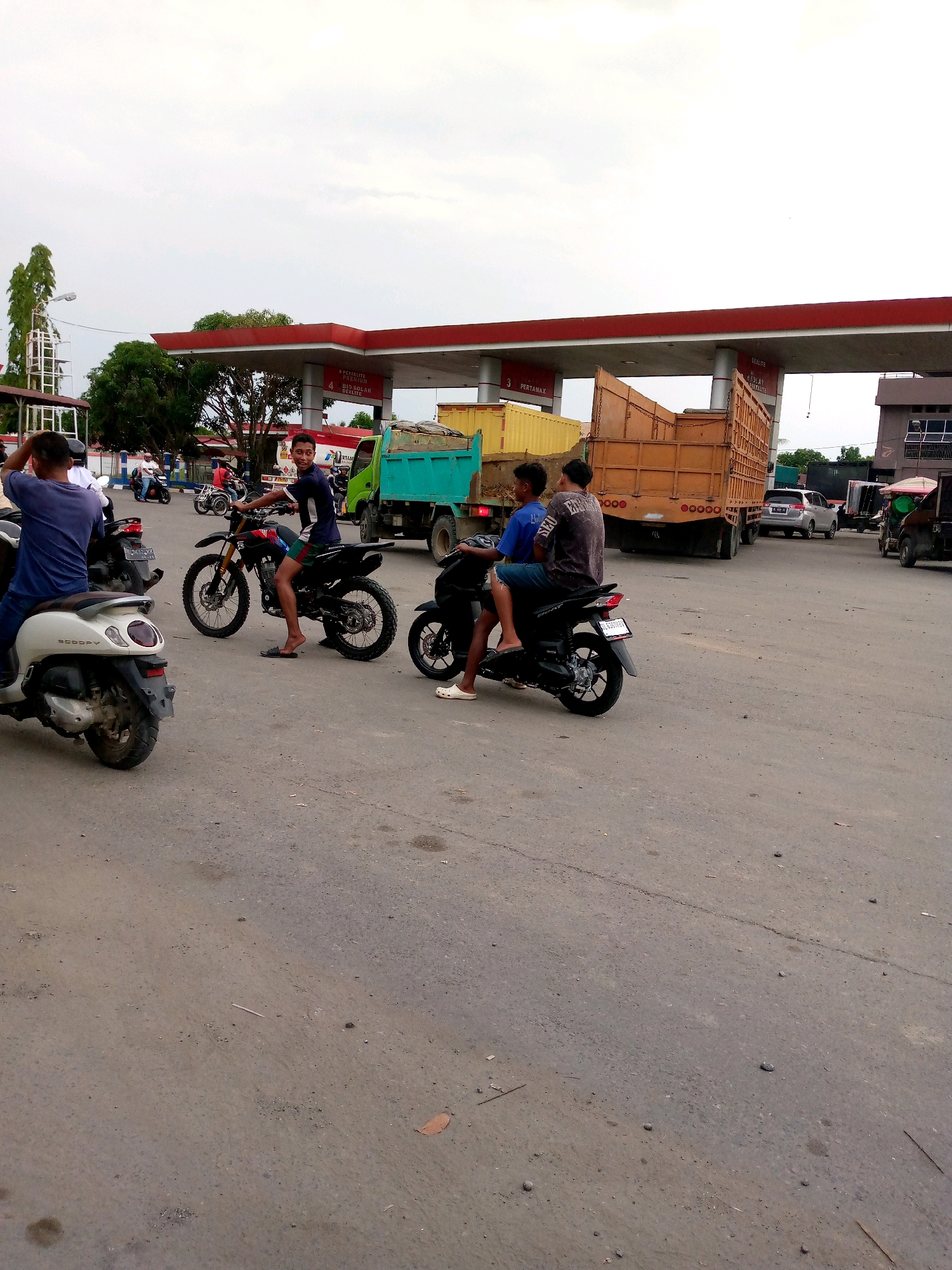 The image depicts a street scene at a gas station in Keudebayu, Indonesia. In the foreground, three motorcycles are visible. The leftmost motorcycle is a white scooter with a person on it facing away from the camera. Next to it, a black dirt bike is ridden by a young man in patterned shorts and a dark t-shirt, looking towards the camera. To the right of the dirt bike, another black motorcycle carries two young men, both wearing t-shirts and shorts.
Behind the motorcycles, a bright green truck is parked. To the right of the green truck is a large, orange flatbed truck. The gas station canopy, painted red with white supports, stretches across the background, displaying signs with Indonesian text, including "PERTAMINA" and names of different fuel types such as "PERTALITE," "PREMIUM," and "SOLAR." Further in the background, a silver car and a darker vehicle are also present. The ground is paved with asphalt. The sky is overcast. The overall environment suggests a busy, everyday scene at a roadside service area.