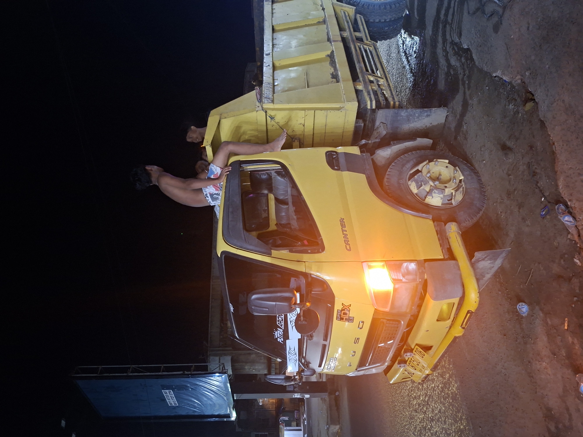 An outdoor, nighttime photograph shows a yellow Mitsubishi Canter Fuso dump truck overturned on its left side on a muddy or unpaved road. The truck's headlights are illuminated, casting a warm orange glow on the immediate surroundings.
Two shirtless individuals, likely young males, are visible interacting with the overturned vehicle. One person, wearing patterned shorts, is positioned with their legs extending upwards from the truck's bed, suggesting they are either inside the cargo area or climbing on it. A second person is partially visible behind them, peering over the edge of the truck bed.
The ground is wet and uneven, covered in mud and dirt, with scattered debris such as plastic bottles and wrappers near the truck's front bumper. In the background, to the left, a dark blue tarp or billboard structure with metal railings is partially visible against the dark night sky.
Visible text on the truck includes "CANTER," "FUSO," and "SUPER HDX 6.6" on the cab. A sticker on the side window reads "PUTRI ARDHANA." The license plate "RL 8750 DB" is clearly seen, along with "06.28," likely an expiry date. The blue structure in the background displays two phone numbers: "0813 7233 4001" and "0813 7120 7752." The scene depicts the aftermath of an incident involving the truck in Keudegeudong, Indonesia.
