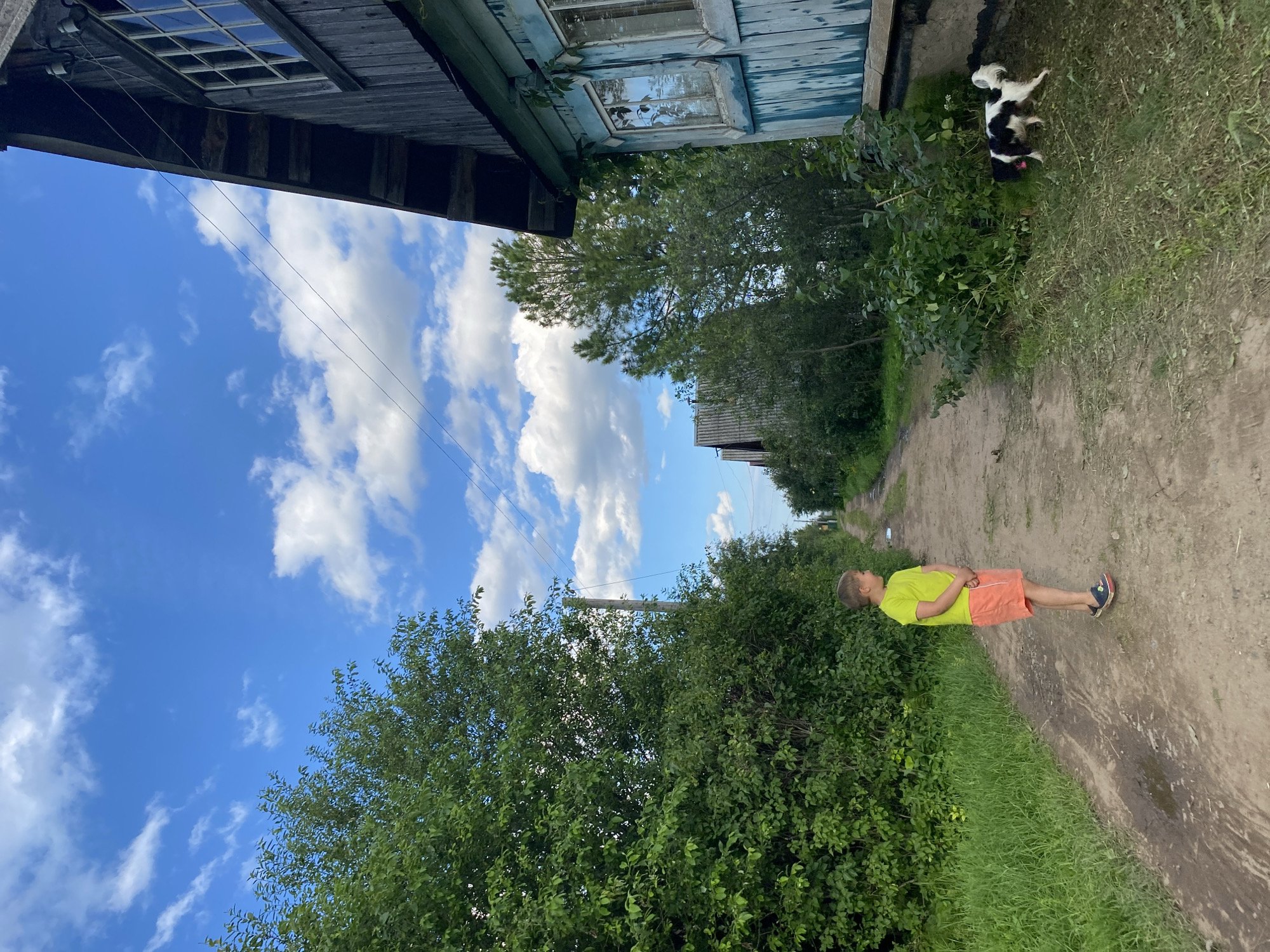 A young boy in a yellow t-shirt and orange shorts lies on a dirt road, his eyes closed and hands clasped over his stomach. He appears to be sleeping or resting in the shade of lush green bushes. The road is flanked by dense vegetation and a weathered, wooden building with peeling blue paint. In the background, a small black and white dog runs across a grassy area next to the building. The sky is a vibrant blue with scattered white clouds, suggesting it is daytime and the weather is pleasant. The scene has a rustic, rural feel, consistent with the location context of Novyi Ural, Russia. There is no visible text in the image.