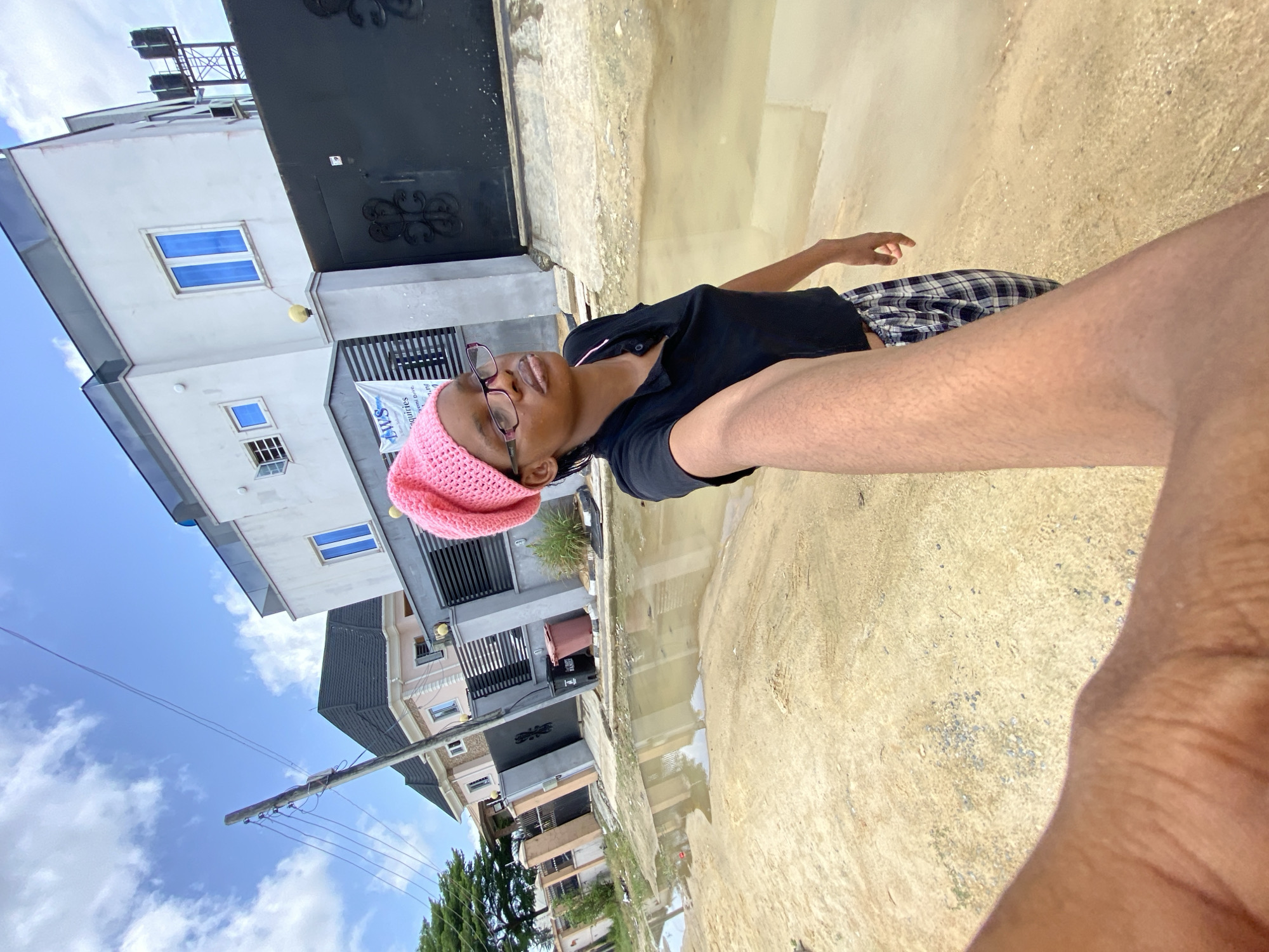 A young woman is depicted in an outdoor setting in Unknown, Unknown. She is wearing a black top, plaid shorts, a pink knit cap, and glasses. The location appears to be a street or walkway adjacent to buildings, with standing water visible in the foreground. In the background, there are residential or commercial buildings under a sky with partly cloudy conditions. Utility poles and wires are also visible.
