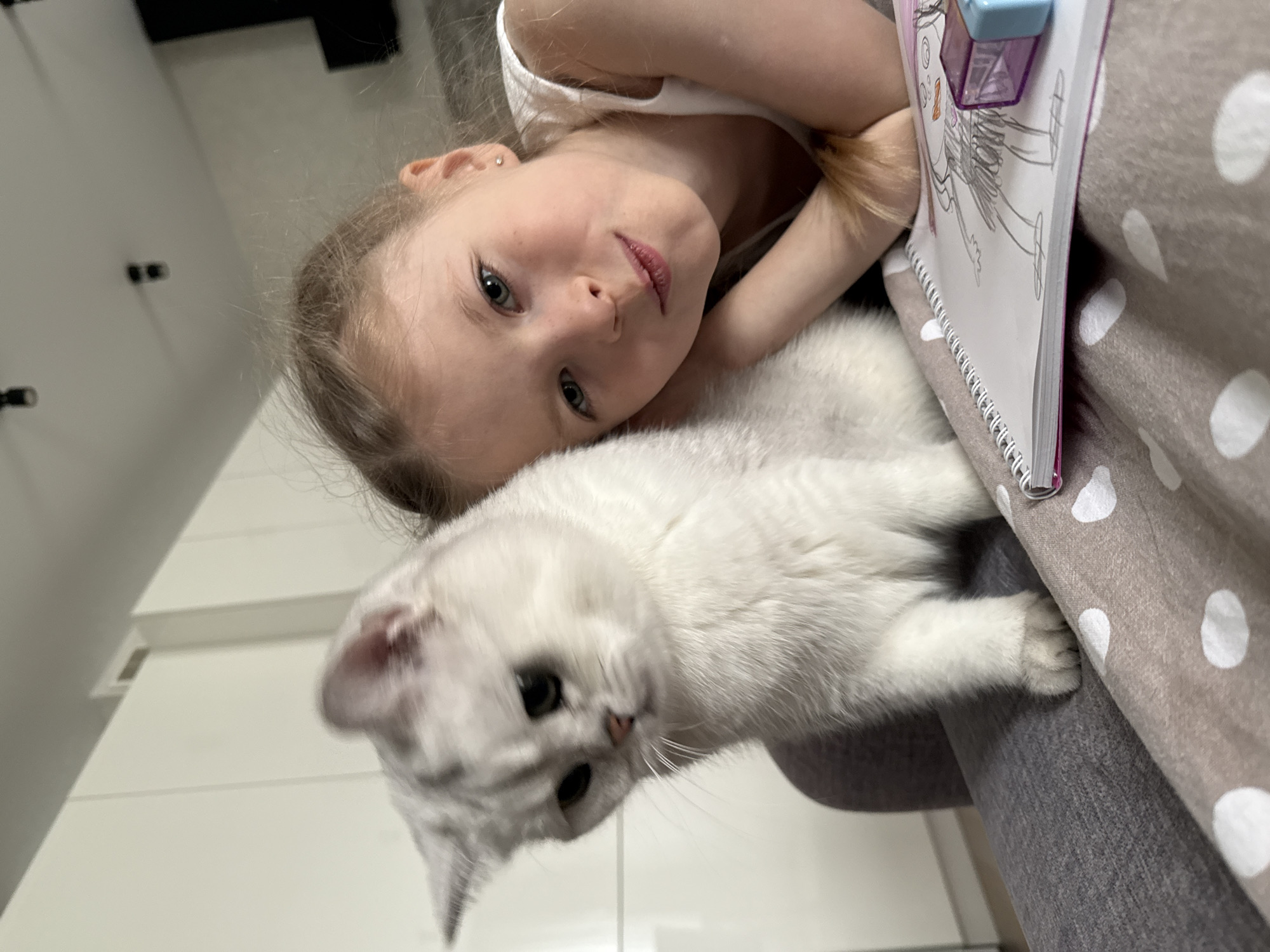 A young girl with fair skin and blonde hair is lying beside a white cat on a surface with a taupe-colored, white polka-dotted fabric. The girl's head is resting on the surface, and she's gazing at the camera. Next to her is a spiral-bound notebook with a drawing and a purple sharpener on top. The cat is looking down and slightly to the left. The background includes a glimpse of a white wall and what appears to be a cabinet. The location is in a residential or indoor setting. The city and country cannot be confirmed.