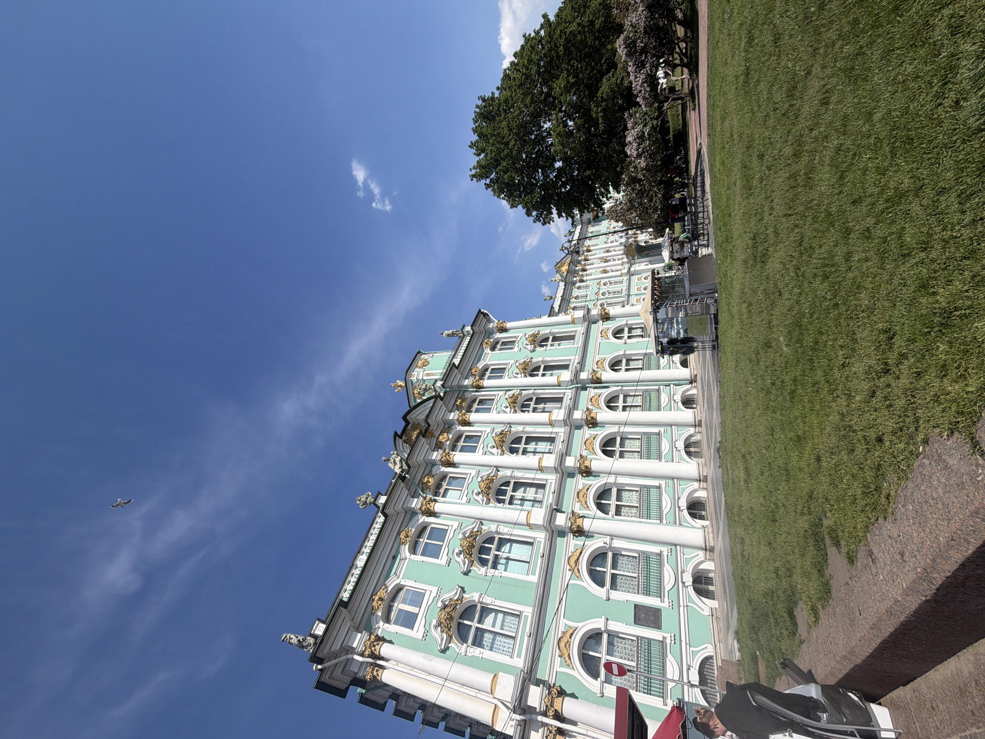 The image shows the Winter Palace in Saint Petersburg, Russia, under a bright blue sky with scattered clouds. The palace, with its distinctive mint-green facade and gold ornamentation, is captured from a low angle, emphasizing its grand architecture. A large, mature tree with dark green foliage stands to the right of the palace, partially obscuring some of its structure. In the foreground, a neatly manicured lawn stretches out, with a granite embankment in the lower left corner. A person is seated on a chair facing away from the camera at the edge of the embankment. To the left of the palace, there's a red awning of a structure. A small, temporary structure with reflective surfaces and some seating is visible in the middle ground, near the palace entrance. Several other people are seen in the distance, milling around in front of the palace and near the trees. A single bird is flying in the sky above the palace.