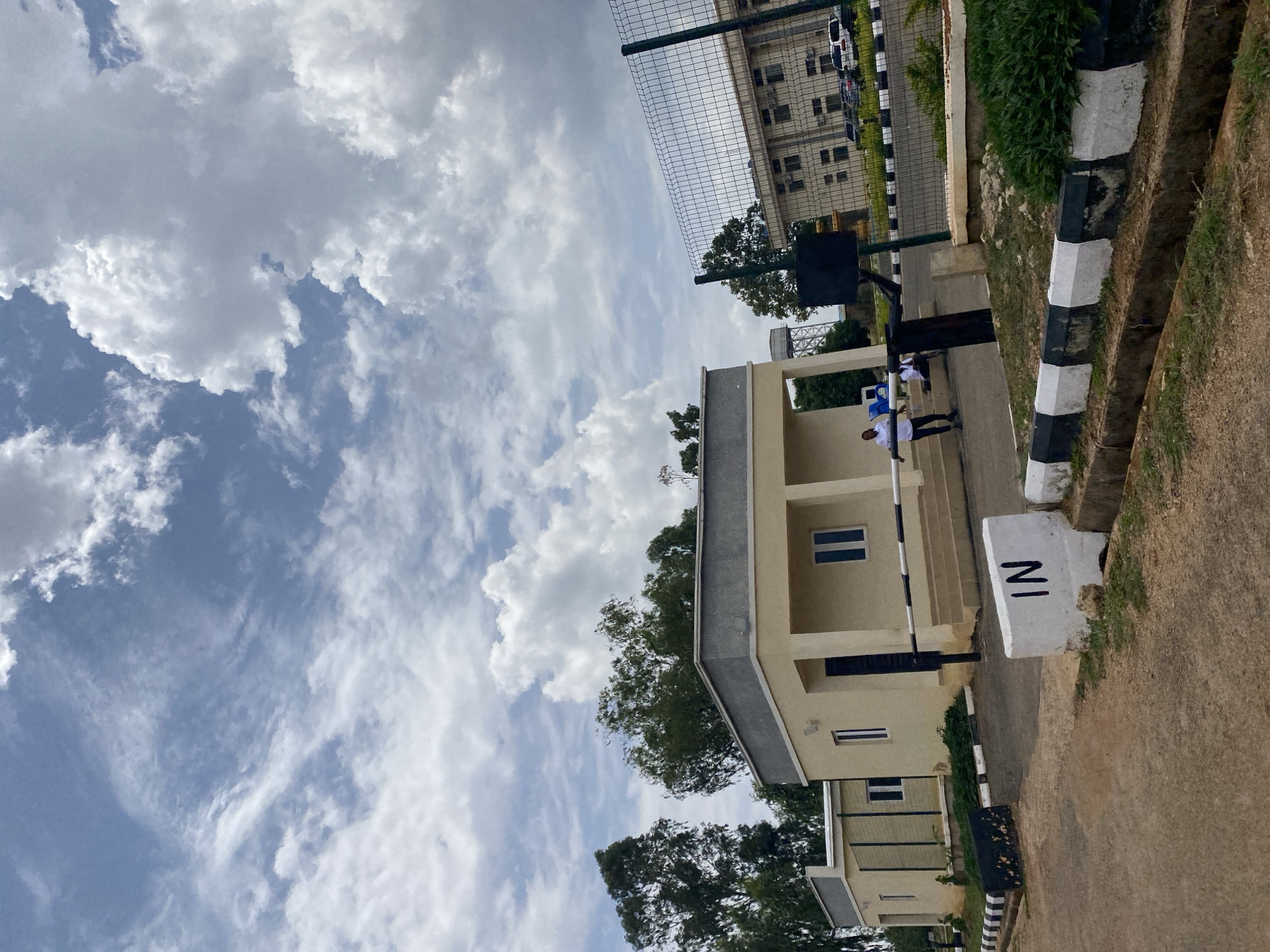 The scene presents a view in Gwong, Nigeria, capturing a beige-colored building with rectangular windows and black window panes, partially visible behind trees. A white pillar with the letters "IN" painted on its side in black is noticeable near the building. Two people, possibly students, are standing close to a stairway attached to the building. The weather is cloudy, with extensive cloud cover dominating the sky. A wire fence is adjacent to a separate, multi-story building with numerous windows and is situated near the edge of the frame. The road is asphalt, and a drainage ditch with alternating black and white vertical stripes is visible along the roadside.