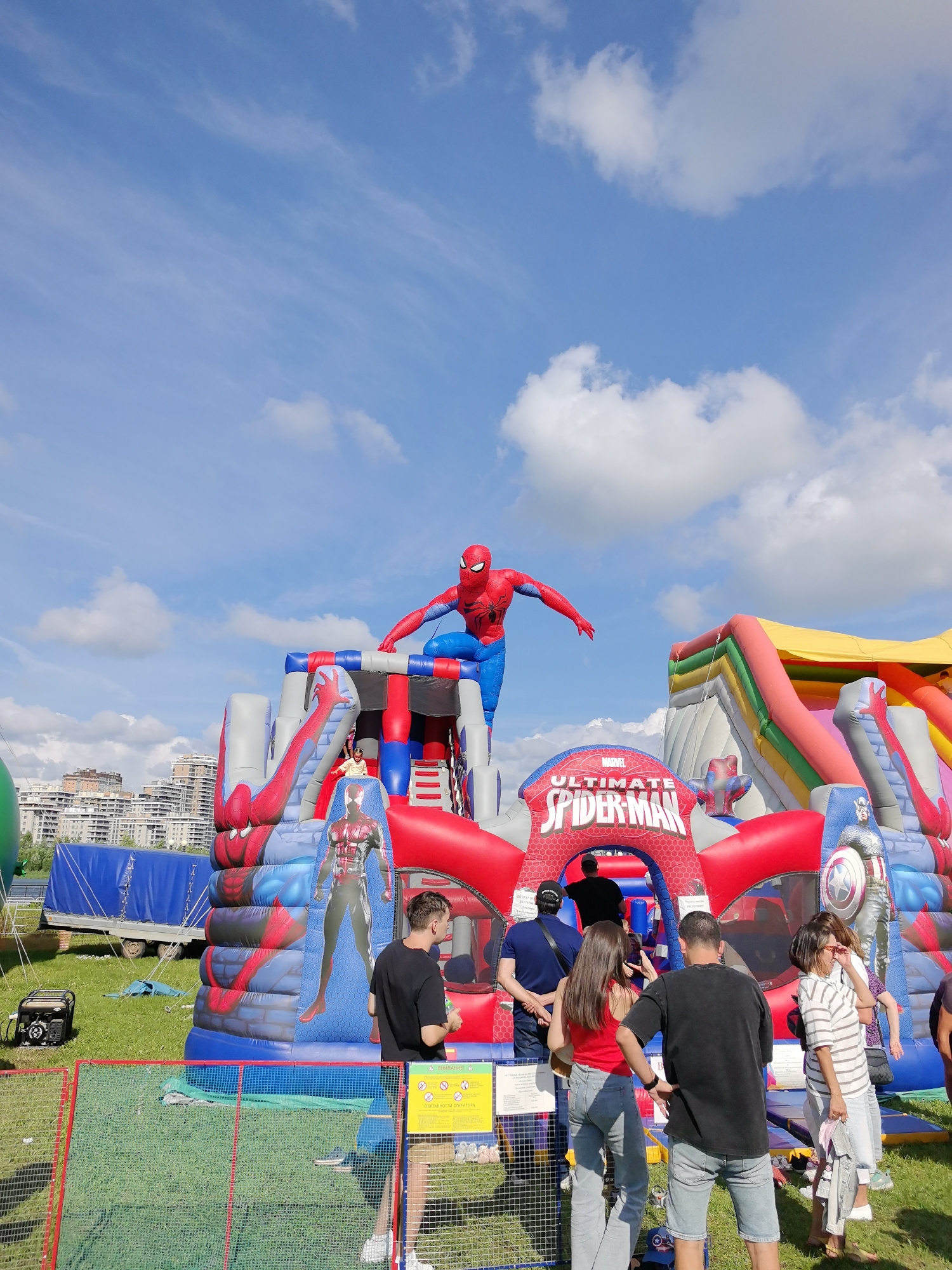 An outdoor, inflatable Spiderman-themed bouncy castle is the central feature of this image, set against a bright blue sky with scattered white clouds. The bouncy castle is large and colorful, with red and blue as the dominant colors, adorned with images of Spiderman and other Marvel characters, including Captain America. A young child in a yellow top can be seen on the bouncy castle's slide. Several people are gathered around the entrance to the bouncy castle, some appearing to be waiting or observing. In the background, there are buildings suggesting an urban environment, and a blue tarp-covered trailer is visible to the left. A portable generator is positioned on the green grass in the foreground, next to a red-fenced area. The scene appears to be an event or a public park. The image was taken in the city of Kazan, Russia.
