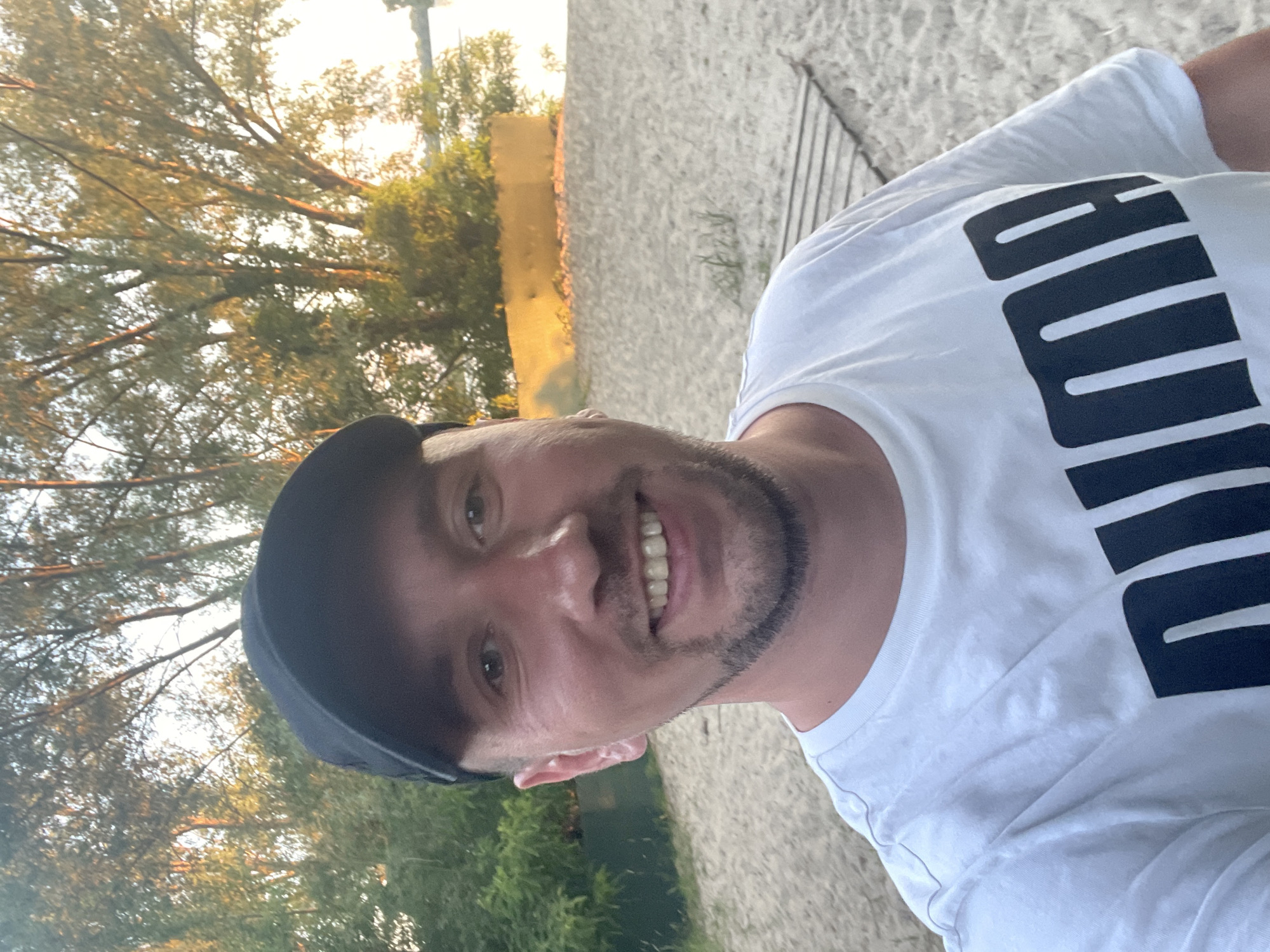A man wearing a black baseball cap and a white Puma t-shirt is taking a selfie on a sandy beach. He has short brown hair, a beard, and is smiling. Behind him, trees and a body of water are visible, suggesting a park or recreation area. The lighting indicates it's late afternoon or early evening. The location is identified as the Hydropark in Kyiv, Ukraine.