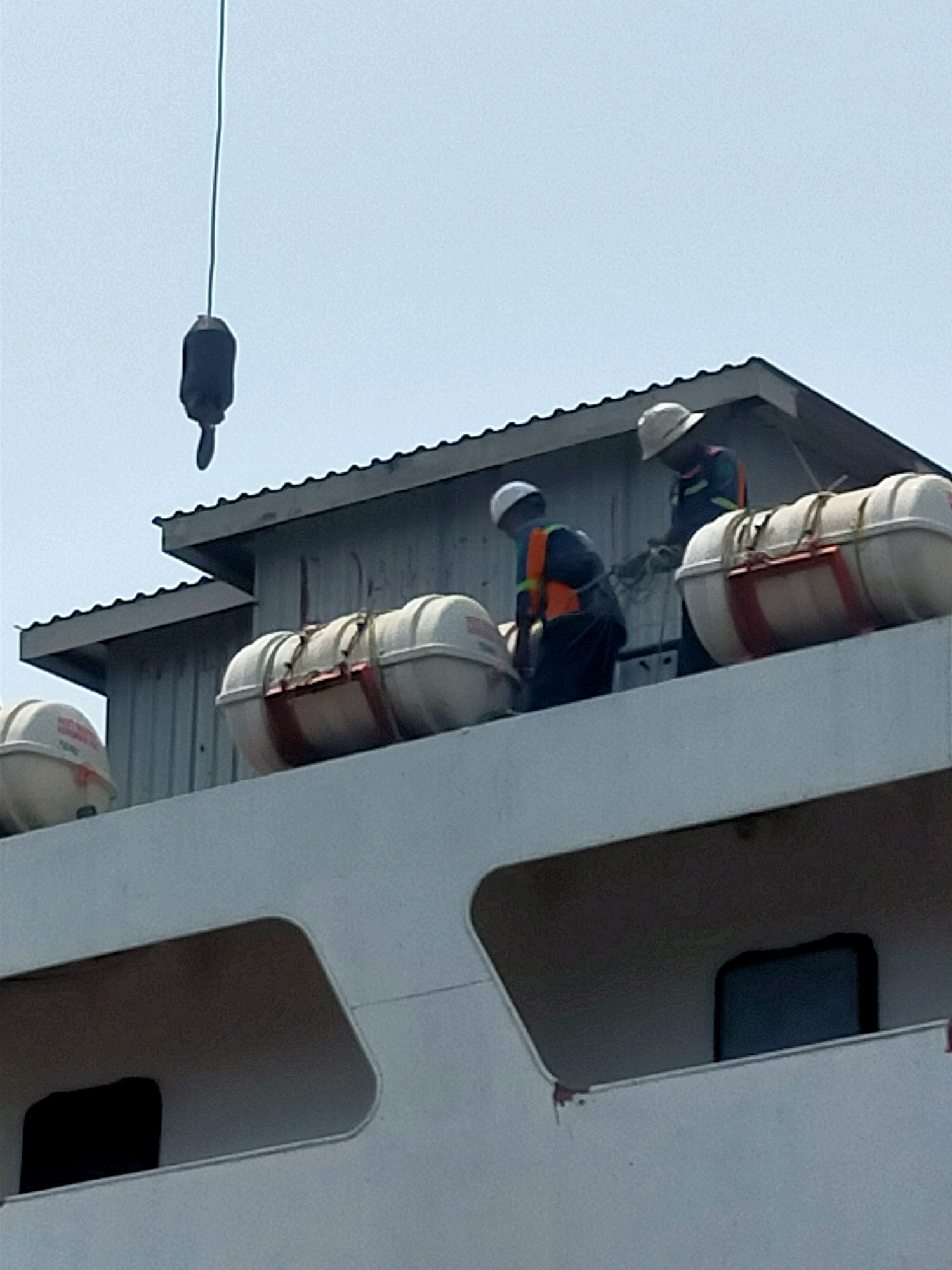 An overhead crane hook is suspended against a hazy sky above a ship. Two workers in safety vests and hard hats are on the deck of the ship, tending to life rafts. One worker is bending over, and the other is standing. The ship's superstructure has a corrugated metal roof. The scene appears to be a harbor in Kamal, Indonesia, likely a working dock given the presence of the crane and ship.
