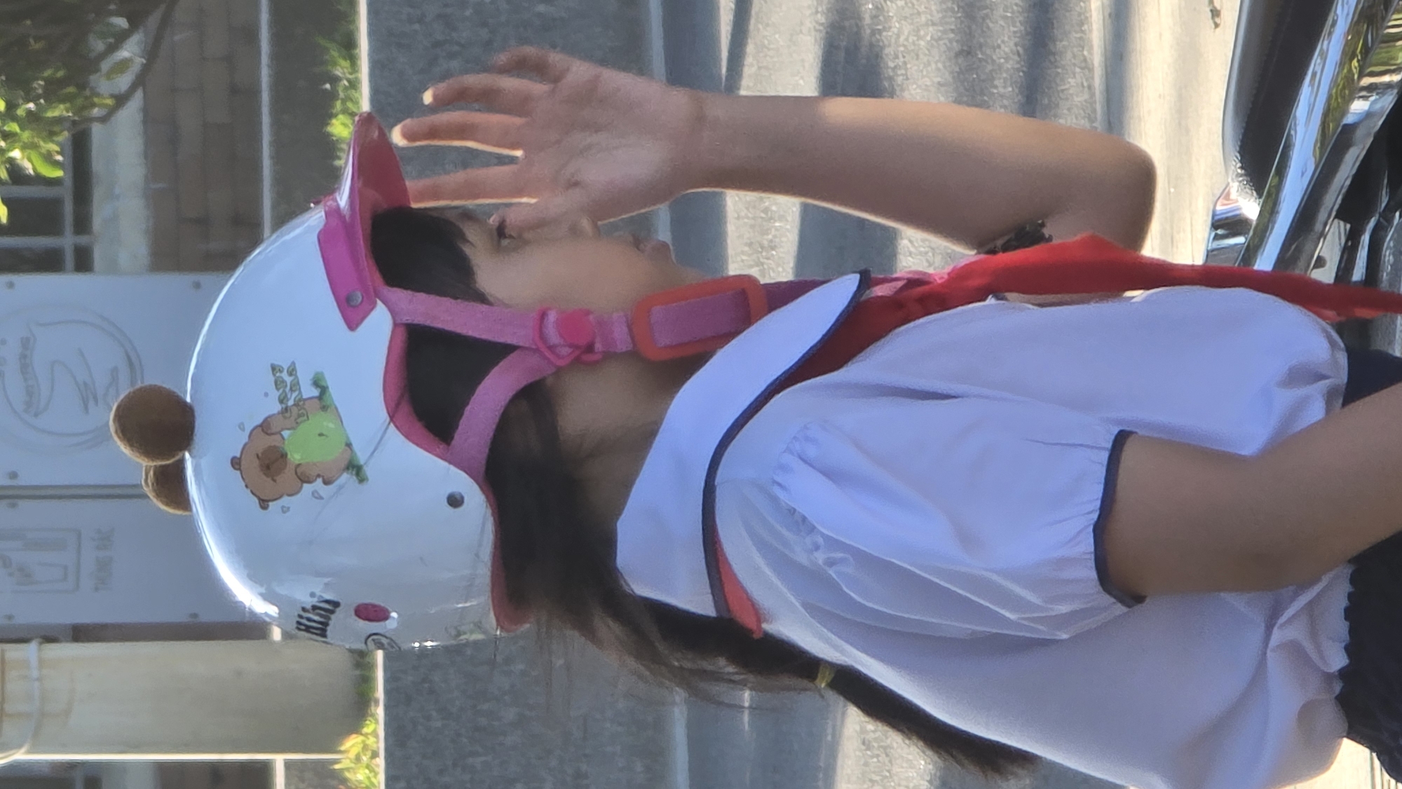 A side view of a child in Nha Trang Ward, Vietnam, wearing a white shirt with a black-trimmed collar and sleeves, along with a red scarf. The child is wearing a white helmet with pink straps and attachments, as well as cartoon-themed stickers. The child's hand is raised and angled to shield the eyes from the sun. The background features a building with window panes, greenery, a sign with a logo, a street, and a motorbike.