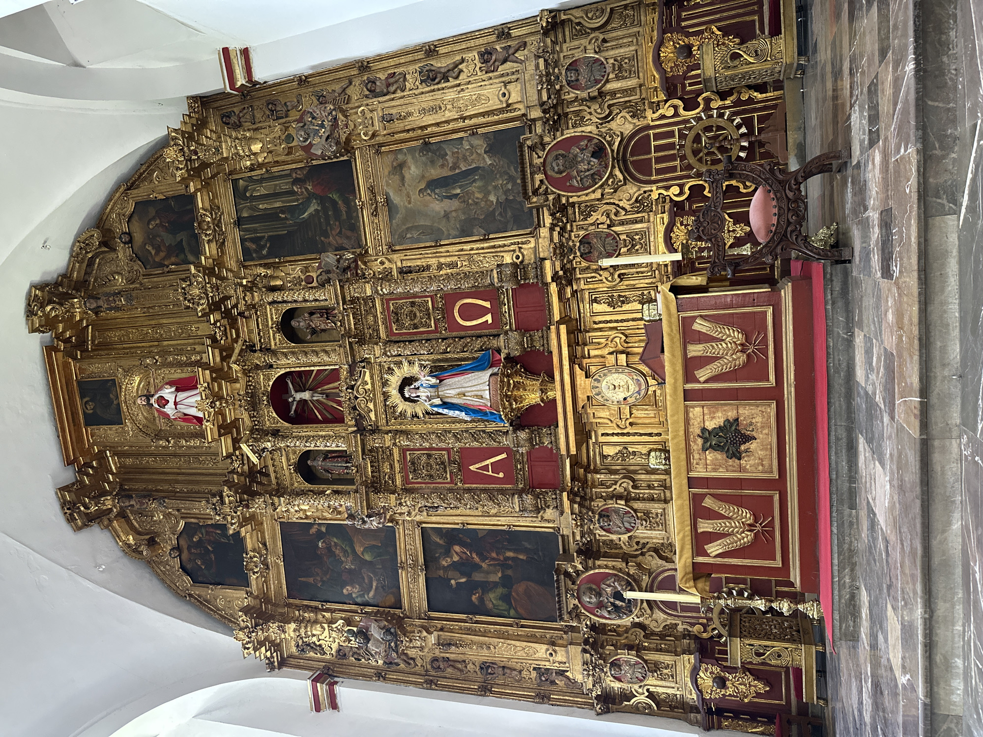 This media file captures a highly ornate, gilded altarpiece, or retablo, located within a church in Mexico City, Mexico. The altarpiece dominates the frame, showcasing a rich Baroque or Churrigueresque style characterized by lavish gold leaf, intricate carvings, and numerous religious figures and paintings.
The structure is multi-tiered and features a complex arrangement of framed paintings, sculptures, and decorative elements. The dominant colors are vibrant gold for the carvings and frames, contrasted with deep red panels. Within these panels are several paintings depicting religious scenes, some with dark backgrounds typical of the era, showcasing figures in various poses and interactions.
Several sculptures are embedded within the altarpiece. Prominently featured are:
* A statue of the Virgin Mary, centrally located, wearing blue and white robes, with a gold halo and base.
* A sculpture of a crucified Christ, positioned above and to the left of the Virgin Mary.
* A figure resembling the Sacred Heart of Jesus, depicted in white robes with a red heart, positioned higher on the left side.
* Other smaller statues of saints and religious figures are integrated throughout the golden framework.
Decorative motifs include numerous cherubs, angels, intricate floral patterns, shell-like designs, and twisted columns, all contributing to the altarpiece's grandeur. On the lower part of the altarpiece, within red panels, are golden depictions of wheat stalks and grapes, common symbols in Christian iconography representing the Eucharist.
Visible text includes two prominent Greek letters: a golden "Ω" (Omega) in a red panel below the Virgin Mary, and a golden "Α" (Alpha) in a red panel above her, symbolizing "the beginning and the end" (Christ).
To the right of the altarpiece, a dark wooden chair with a pink velvet cushion is visible, featuring ornate carvings and a circular decorative element. The church interior also shows a white vaulted ceiling and a floor made of large, polished grey marble tiles. No specific activity or event is taking place; the scene presents a static view of this significant religious artwork, conveying a sense of solemnity and spiritual devotion.
