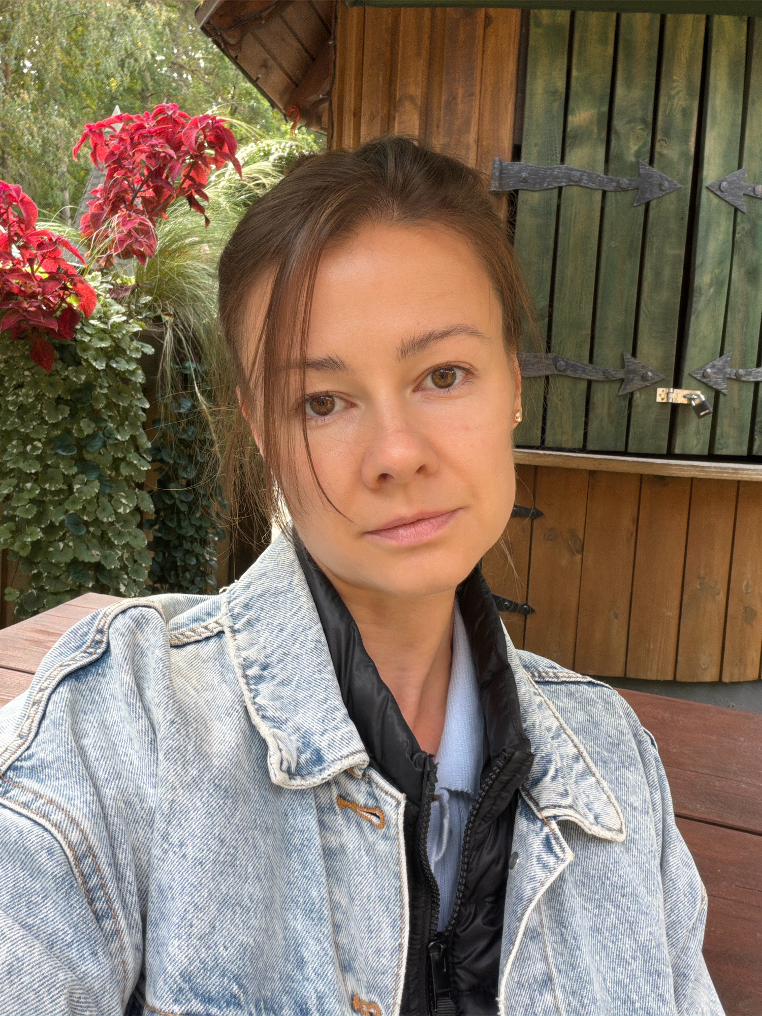 A medium shot photograph shows a woman from the shoulders up, sitting at a wooden table outside. She has brown hair pulled back in a messy bun and is wearing a denim jacket over a puffer vest and a light blue shirt. She is looking directly at the camera with a neutral expression. Behind her, there is a wooden structure with green doors that appear to be ornately decorated with metal hinges. To the left of the woman is a planter with lush green foliage and bright red flowers. The overall setting appears to be a garden or an outdoor seating area, possibly at a park or a restaurant. The style of the wooden structure and the presence of decorative hinges suggest it could be a historical or themed building. The image captures a natural lighting condition, with the sun casting subtle shadows. The location is identified as Tērvete, Latvia, which is known for its natural beauty and popular tourist attractions like the Tērvete Nature Park.
