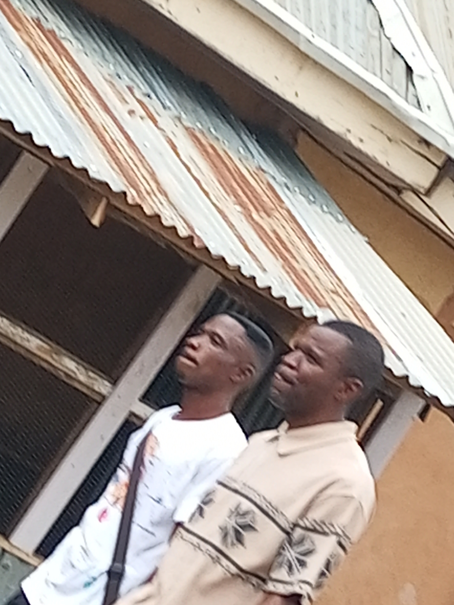 The image captures two young African men in an outdoor setting, possibly in Tempure, Nigeria. They are standing near a weathered corrugated metal roof, which shows signs of rust. One man on the left is wearing a white shirt with a dark strap across his chest. The man on the right is wearing a light-colored collared shirt with a patterned design on the sleeves and chest. Both men appear to be looking upwards and slightly to their right, suggesting they are observing something beyond the frame. The background includes elements of a building, with wooden structures and a window with a dark mesh screen. The lighting suggests it is daytime.