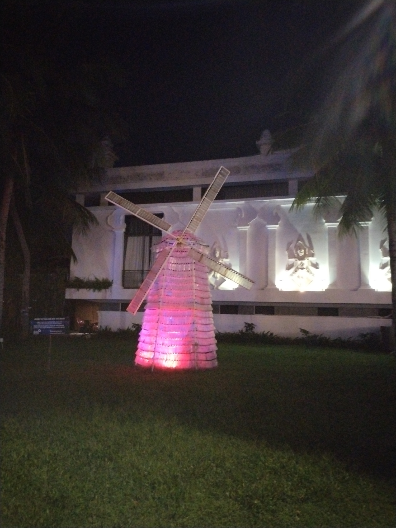 The image depicts a nighttime scene in Bac Nha Trang Ward, Vietnam, featuring a makeshift windmill constructed from translucent plastic bottles, illuminated with pink and red lighting. The windmill stands on a green lawn between two palm trees. Behind the windmill is a white building with ornate columns and sculptures. A sign is visible in the lower left, partially obscured by darkness.