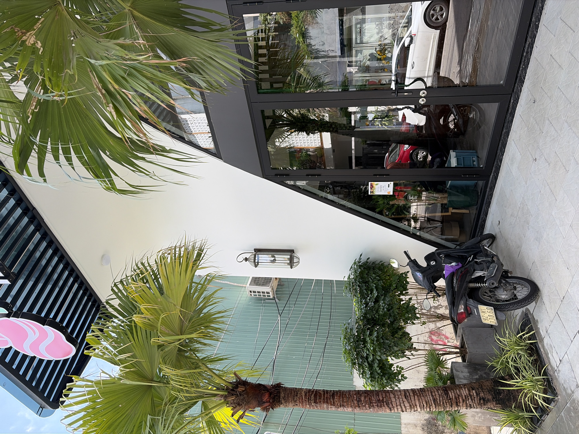 The image captures the exterior of a modern building in the city of Da Nang, Vietnam. The building features a striking white, angular facade that contrasts with dark-framed glass doors and windows. A large palm tree stands prominently in the foreground to the left, its fronds reaching towards the sky. Adjacent to the building's entrance, a dark-colored motorcycle is parked on a paved walkway, with its license plate visible. Lush green bushes and potted plants are situated near the building's base, adding a touch of nature to the urban environment. Reflections of a street with cars are visible in the glass doors. The overall scene suggests a commercial establishment, likely a cafe or shop, with a contemporary architectural style.