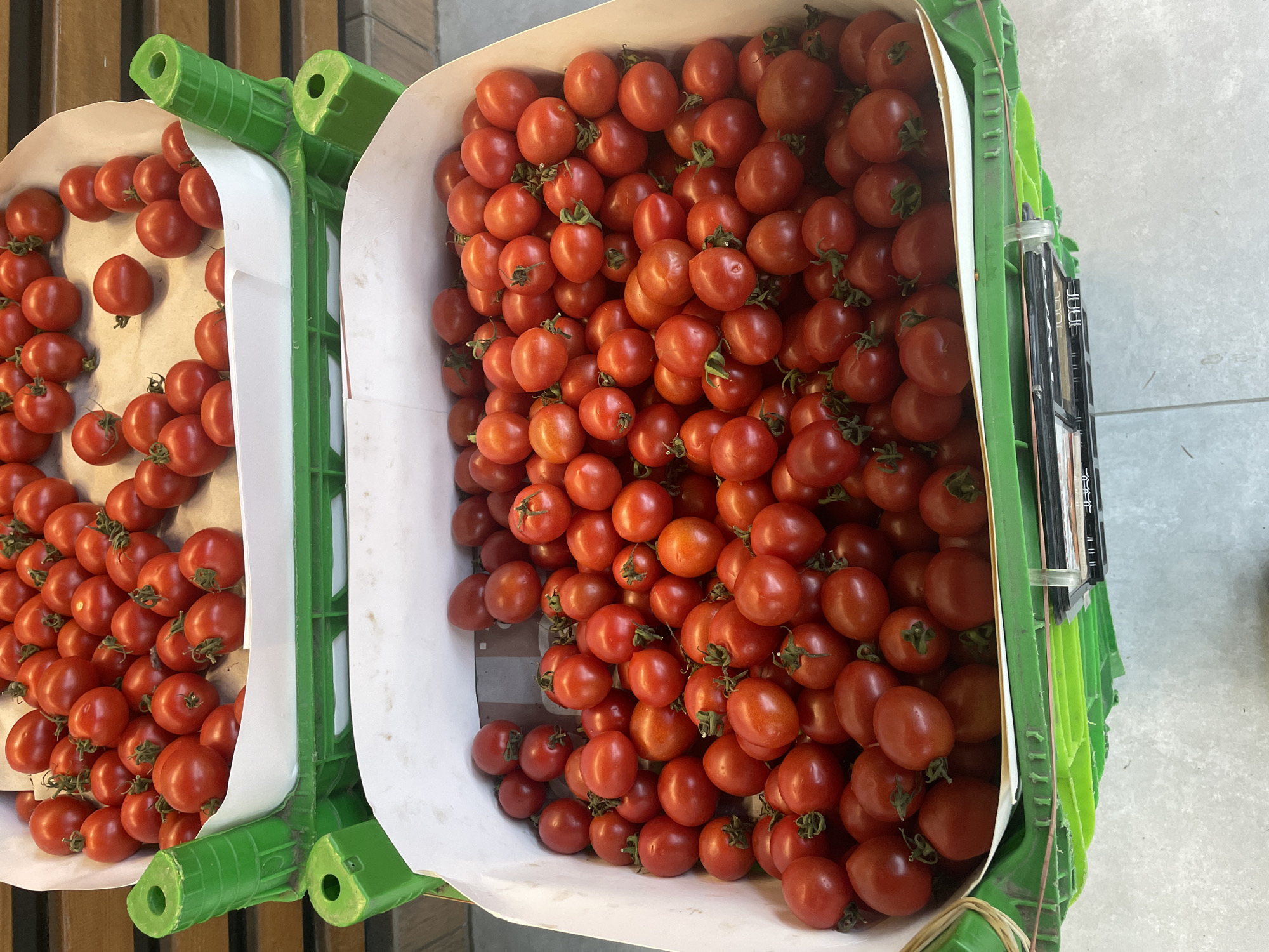 The image shows two green plastic crates filled with ripe red cherry tomatoes. The crates are stacked, with the top crate partially visible and the bottom crate in full view. The tomatoes are small, round, and appear fresh. The crates are positioned on a tiled floor, suggesting a market or grocery store setting. The lighting is bright, illuminating the vibrant color of the tomatoes. The background is blurred, with hints of wooden shelving. The scene does not contain any people or identifiable landmarks of Kadıköy, Turkey.