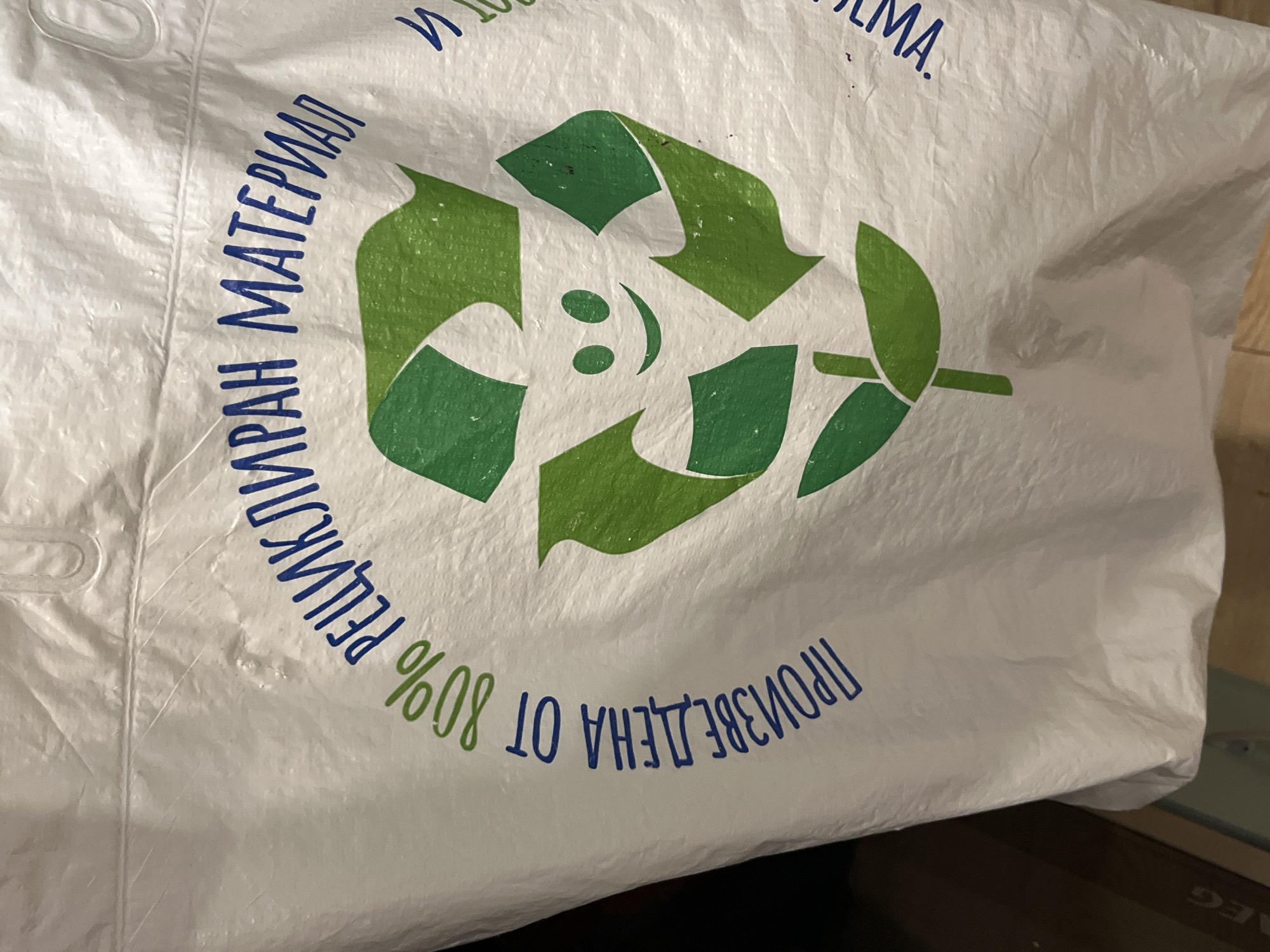 The image displays a close-up of a crinkled, white woven polypropylene bag, indicative of a reusable shopping bag. The bag is adorned with text in Bulgarian Cyrillic and a stylized recycling logo.
In the upper central area, blue Cyrillic text arcs across the bag, reading "РЕЦИКЛИРАН МАТЕРИАЛ" (Recycled Material). To the left, following a descending arc, green text states "80%", followed by blue text "ПРОИЗВЕДЕНА ОТ" (Made from). This collectively indicates the bag is "Made from 80% Recycled Material."
Dominating the center of the bag is a two-toned green recycling symbol, composed of three chasing arrows arranged circularly. Uniquely, the negative space within the symbol forms a smiling face with two dots for eyes and a curved line for a mouth. The lower portion of the symbol extends into a green stem with two small leaves, resembling a sprout. Partially visible at the top are clear plastic loop handles for the bag.
The background is out of focus. On the lower left, a dark brown object, possibly furniture or an appliance, displays the partial white lettering "AEG." The lower right reveals a light brown surface, likely a wooden floor. The scene is indoors.
The Bulgarian text confirms the image's location in Bulgaria, specifically in the city of Pazardzhik.