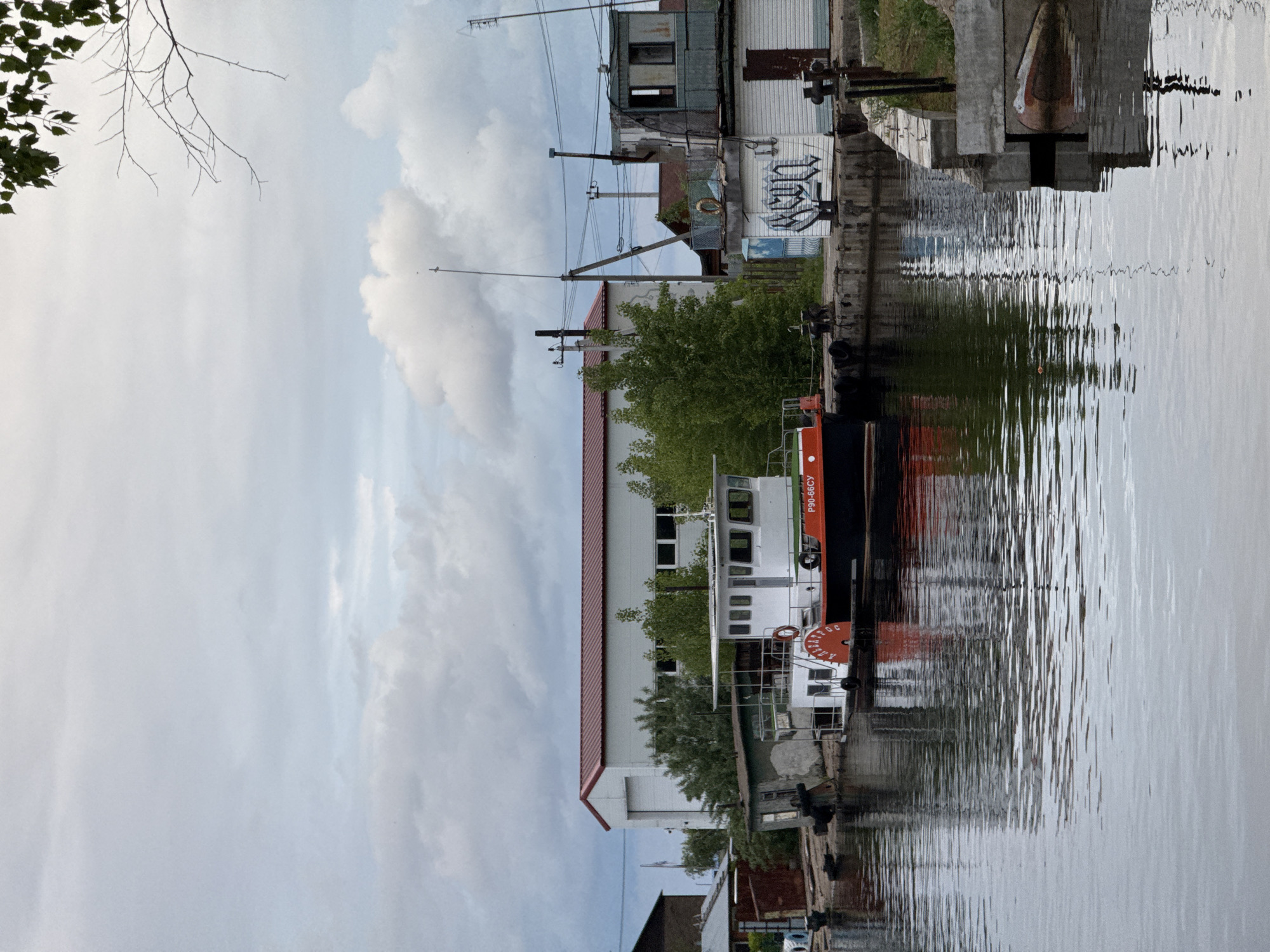 The image shows a waterfront scene in what is likely Chaikovsky, Russia, given the Cyrillic lettering on the boat. The foreground is dominated by the calm water of a river or harbor. A boat with white and orange paint, labeled "P90-56CY" in Cyrillic, is docked alongside a concrete quay. The reflections of the boat, buildings, and greenery are visible in the water. On the quay, there are several buildings with varied architecture. Some appear to be older industrial buildings with visible signs of wear, while others look more modern. The sky is overcast with grey clouds, suggesting a cloudy or possibly rainy day. There are trees and other vegetation intermixed with the buildings, adding a touch of green to the scene. Utility poles with wires are also visible, contributing to the urban landscape. The image is oriented sideways, requiring the viewer to rotate it for a proper perspective.