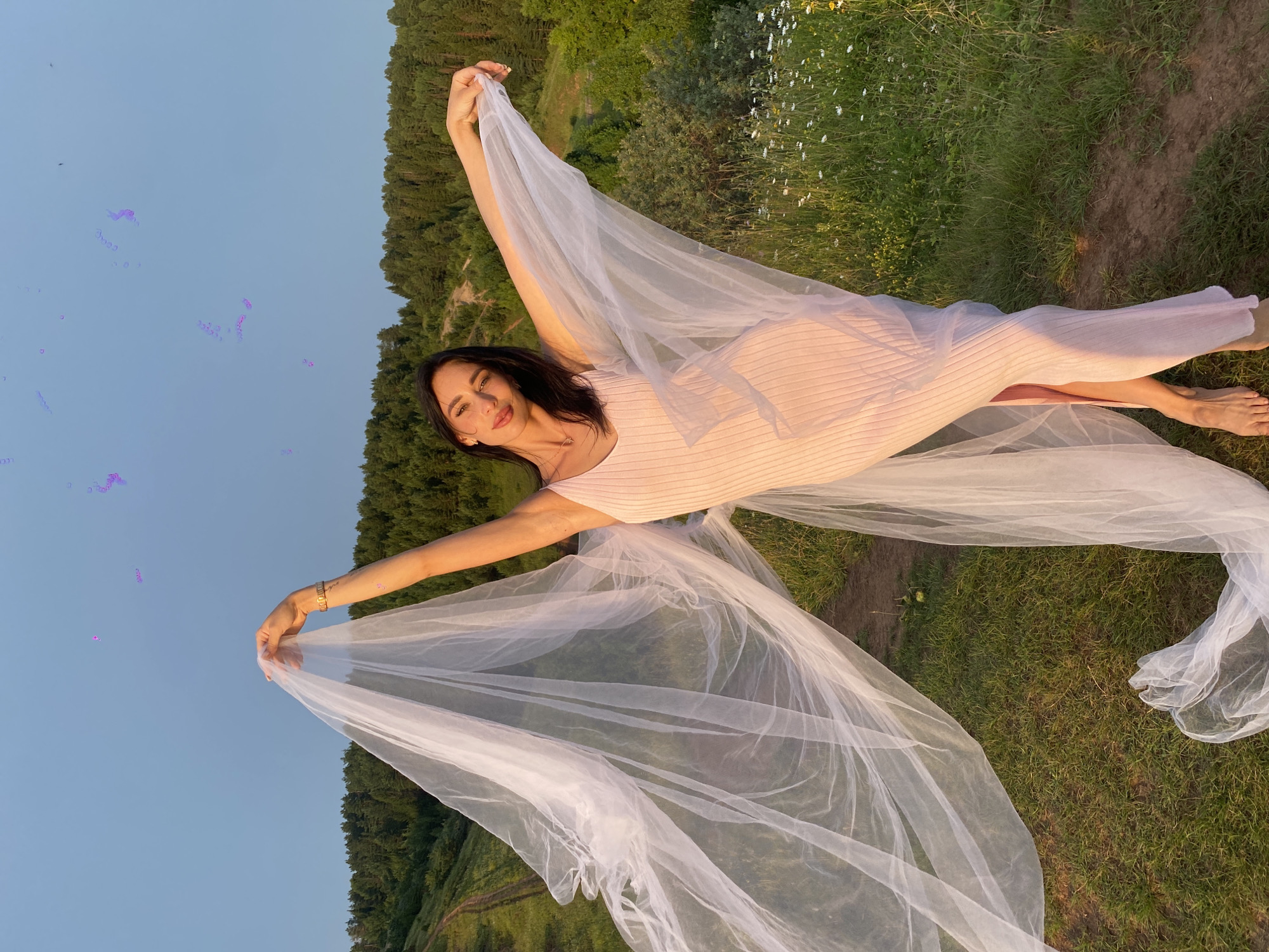 A woman with dark hair and fair skin stands in a grassy field with trees in the background. She is wearing a light pink, ribbed dress and holding a sheer white veil that billows around her like wings. The sky is a clear blue with some faint purple streaks. The ground is covered in grass and wildflowers, and a dirt path winds through the scene. The overall impression is one of ethereal beauty and freedom. The image is likely a photograph taken in Olkhovka, Russia, given the natural landscape.