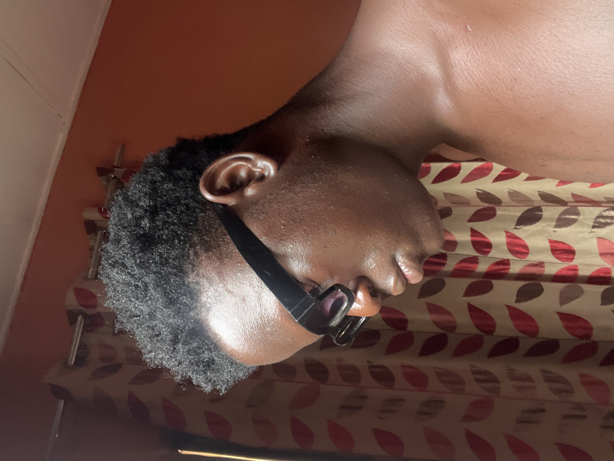 The image shows a close-up of a person's head and upper chest, wearing glasses, in an indoor setting. The person has dark skin and short, curly black hair. They are lying down on a surface covered with a patterned fabric of leaves in red and brown. The wall behind the person is painted in orange. The location is presumably a residential setting. There is no other evidence to indicate that this is in the city of Mosafejaw, Nigeria.