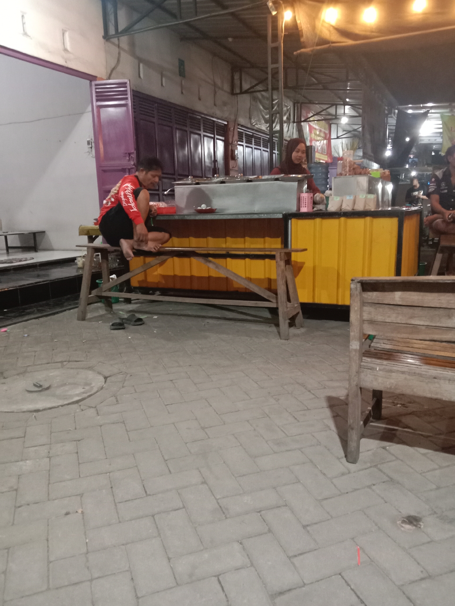 At night, in front of a building with a corrugated metal roof, a man in a red shirt and black shorts sits on a wooden bench at a food stall. He is facing a counter with several dishes. To his right, a woman wearing a hijab and a red top serves customers. The food stall is decorated with yellow corrugated metal. The ground is paved with interlocking gray bricks. A wooden bench is located in the foreground. A person is seated on a stool to the right, partially visible. The scene suggests a local eatery or market in Ketapang, Indonesia.